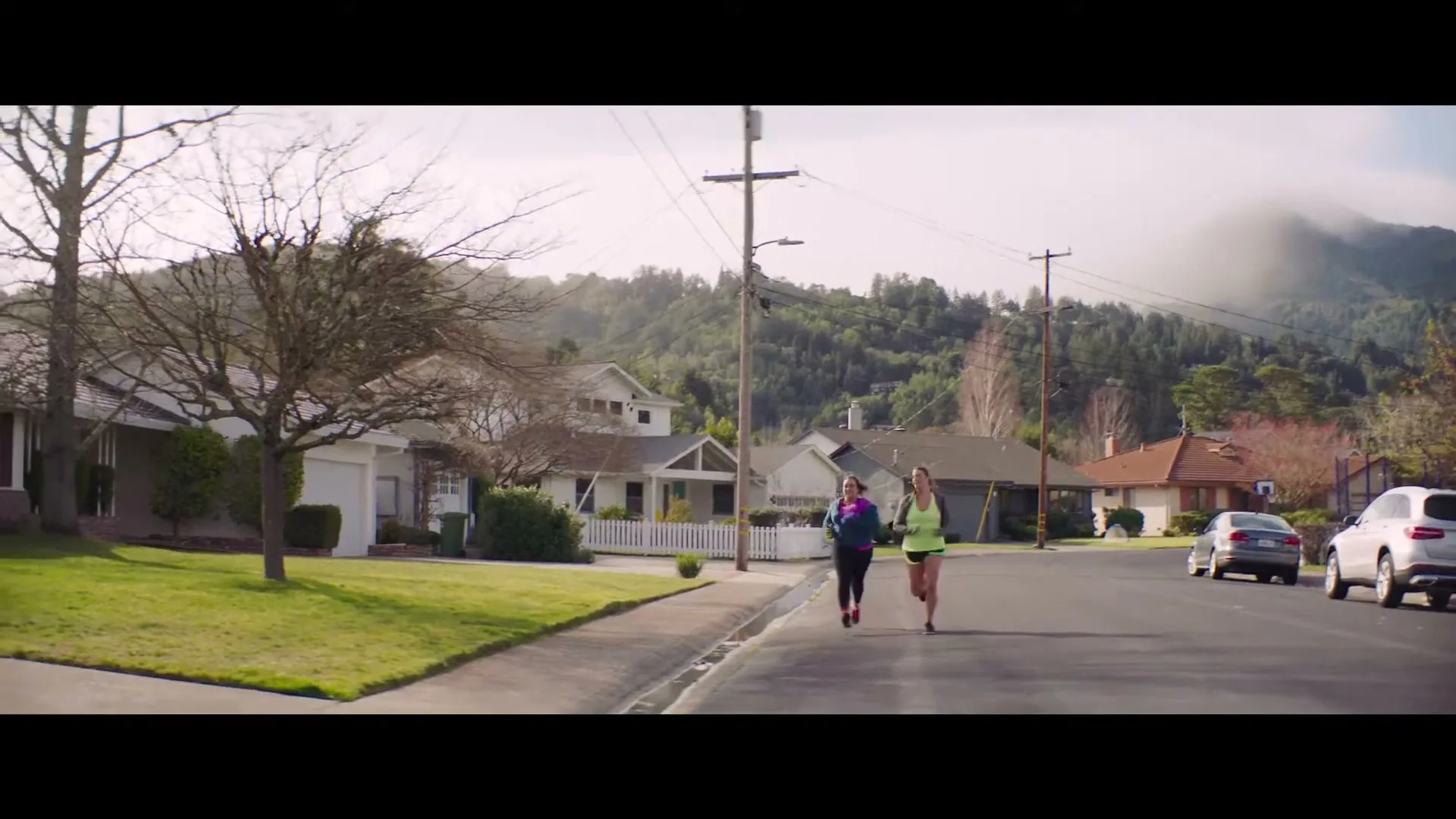 Two women in athletic wear run down a suburban street with houses and trees on either side, under a bright sky with distant mountains.