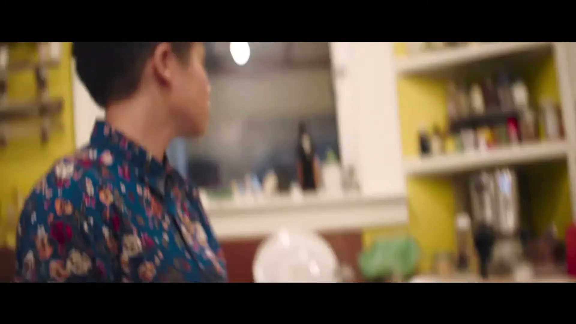 A young person with short dark hair and a floral shirt leans over a stove, then serves noodles onto plates in a kitchen.