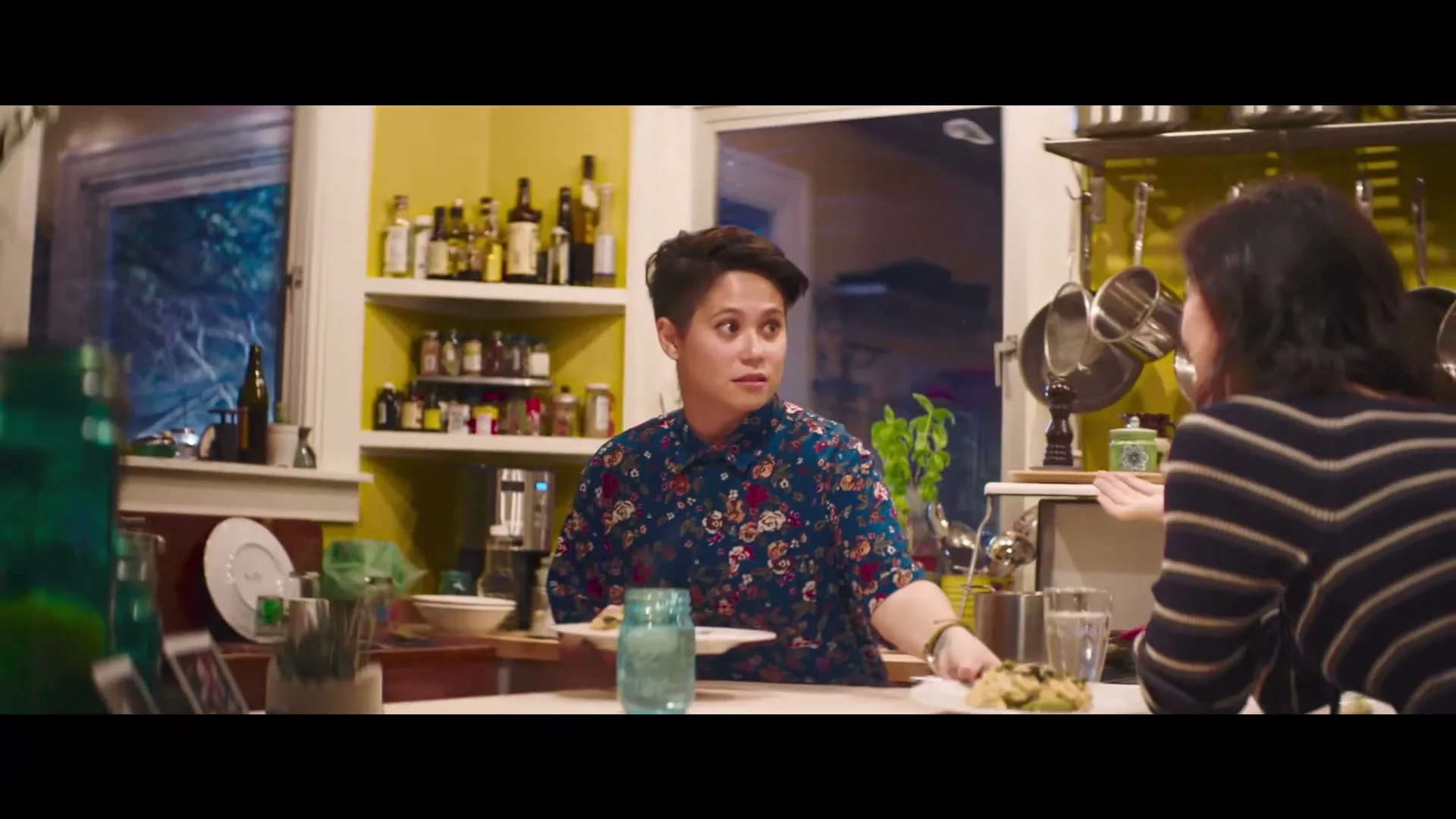 A person with short hair in a floral shirt serves food to another person at a kitchen counter, looking surprised.