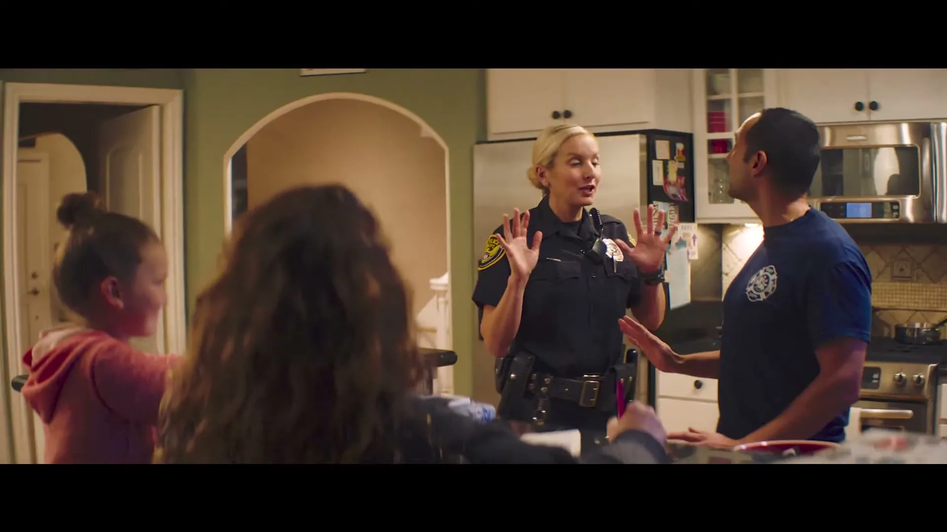 A female police officer and a male firefighter talk in a kitchen, while two children are seen from behind in the foreground.