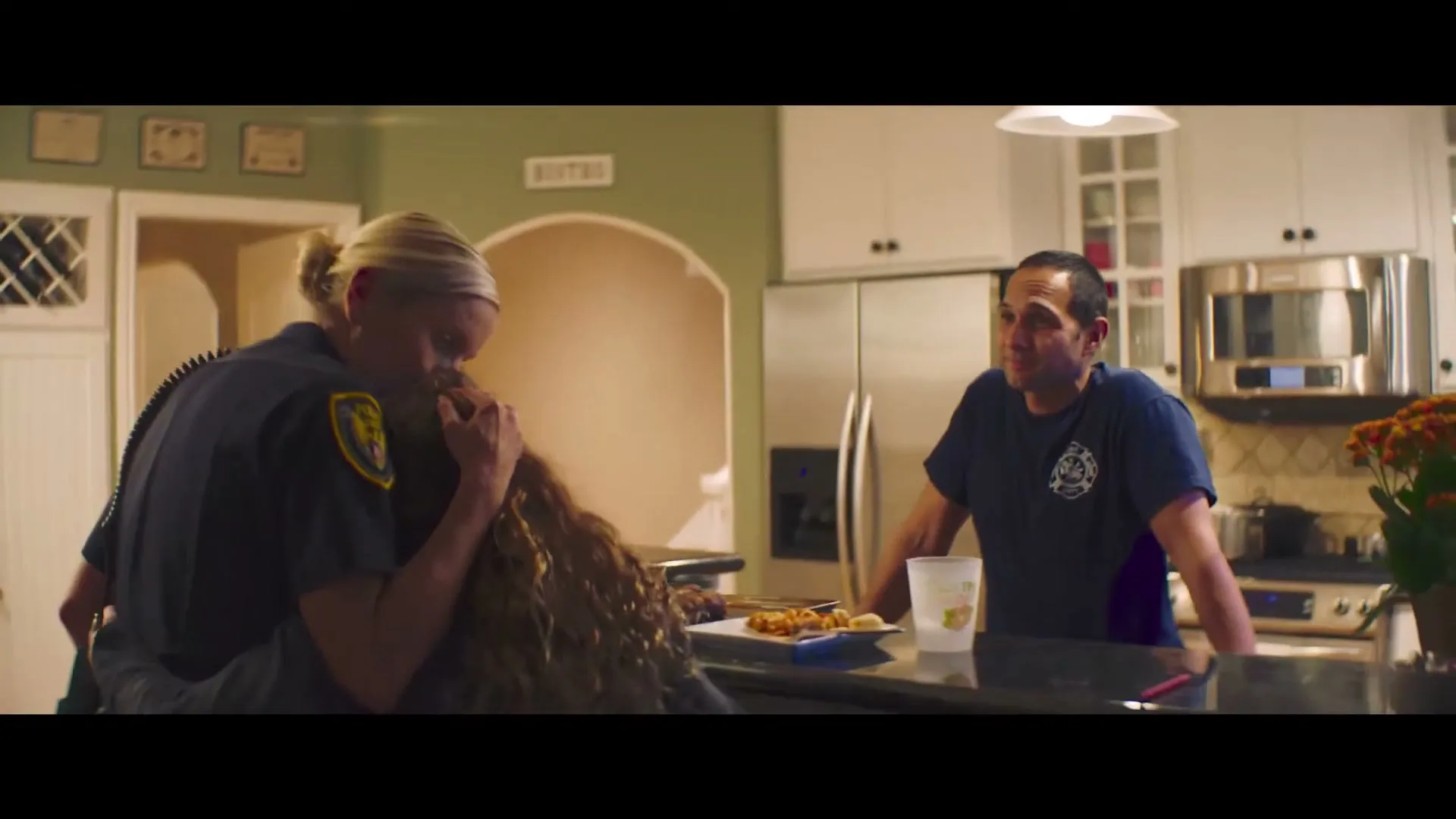 A female police officer hugs a young girl with curly hair in a kitchen, while a male firefighter watches them from across the counter.