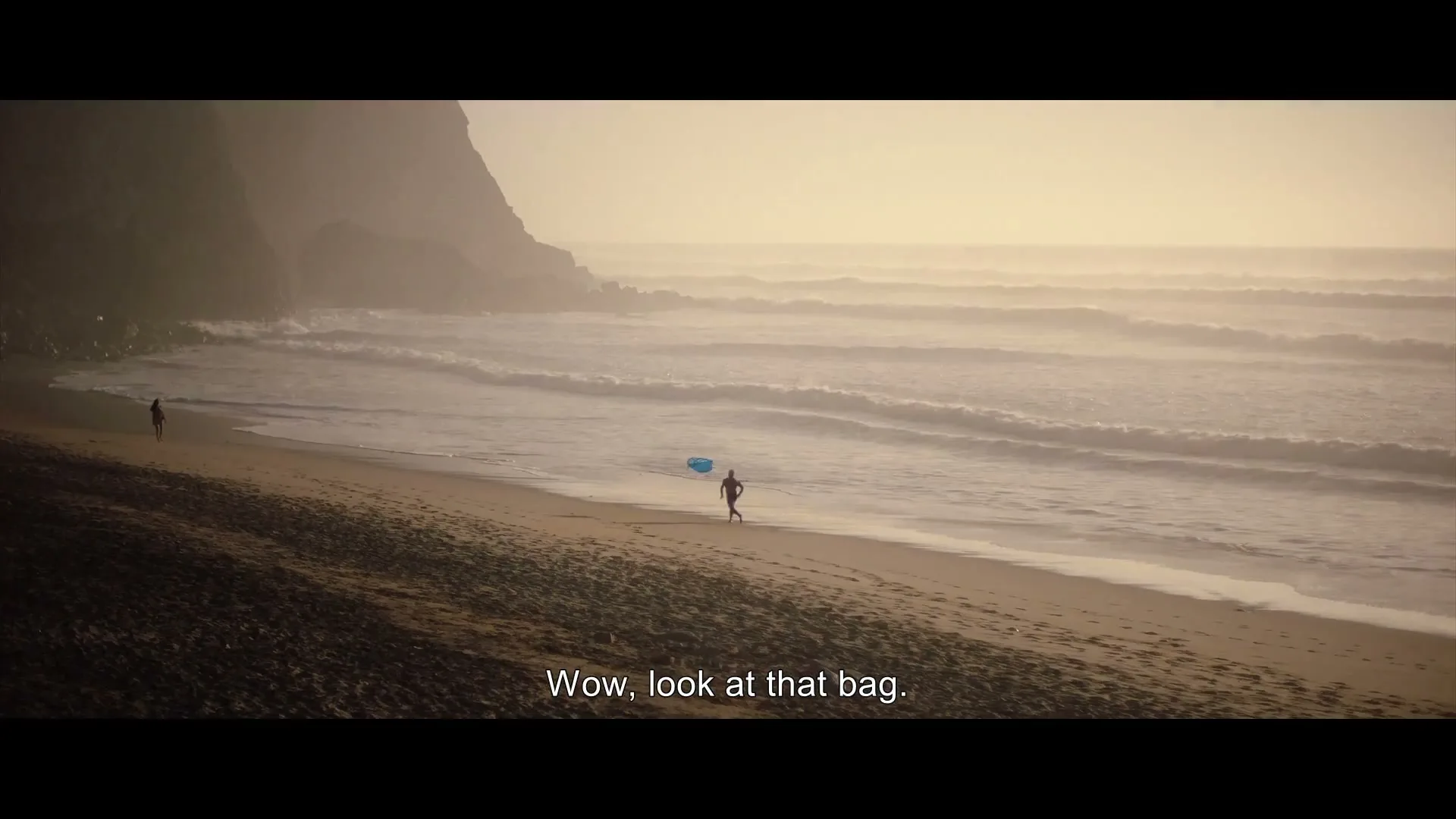 A person in dark clothing runs along a sandy beach carrying a blue bag, with another person in the distance and waves crashing on the shore under a hazy sky.