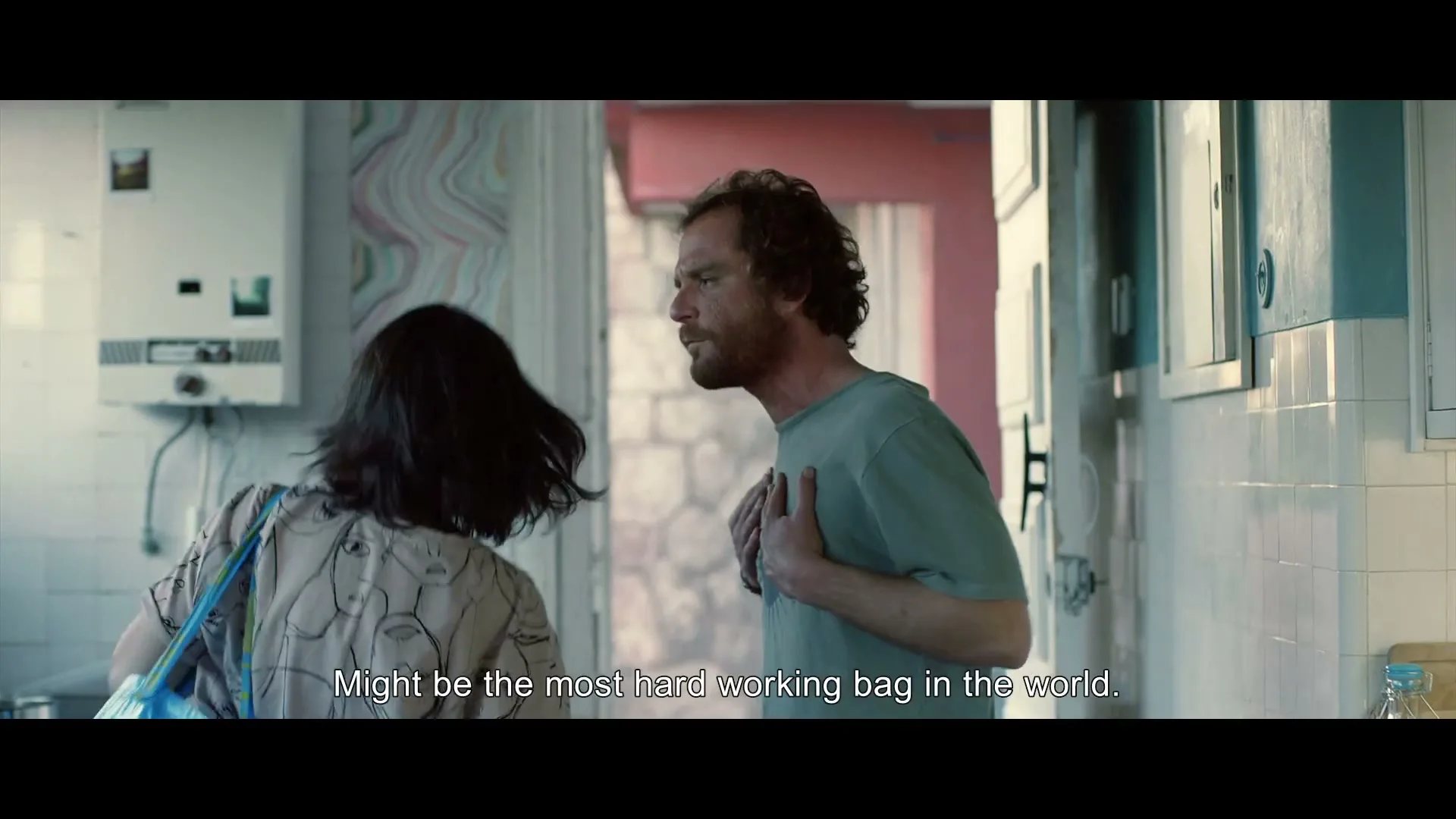 A man with a beard and a woman with dark hair are in a kitchen, the man is talking to the woman who is holding a blue bag.