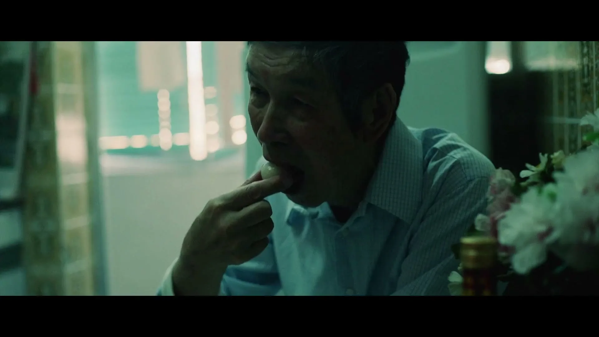 An elderly Asian man in a light blue shirt eats a small white food item with his fingers in a dimly lit room.