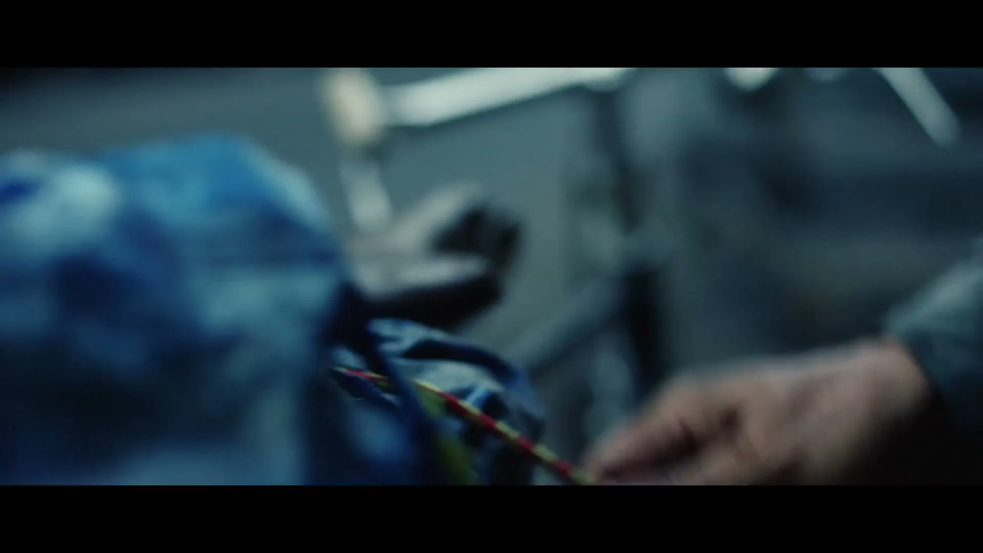 A man's hands are shown in a medium shot, tying a colorful rope around a blue bag in a dimly lit, industrial setting.