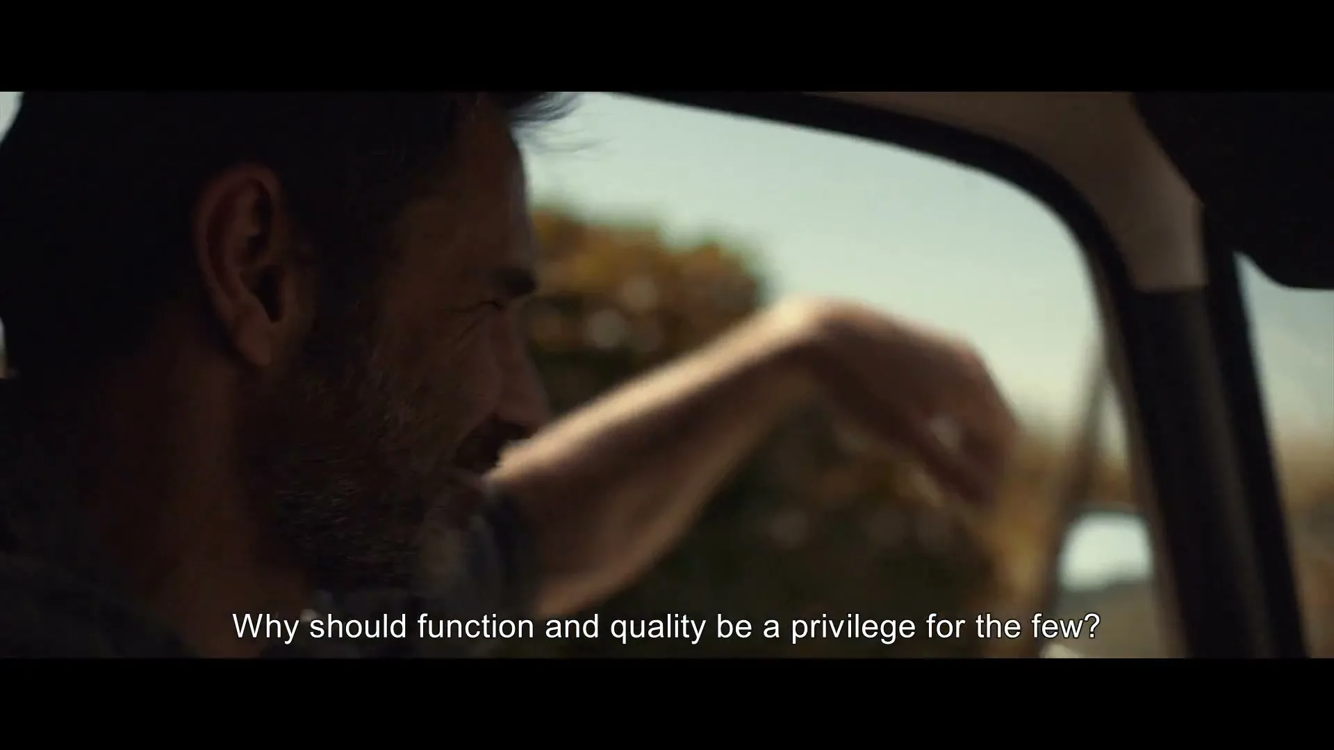 A man with a beard drives a car, looking out the window with a slight smile, as a woman's arm is visible outside the car.