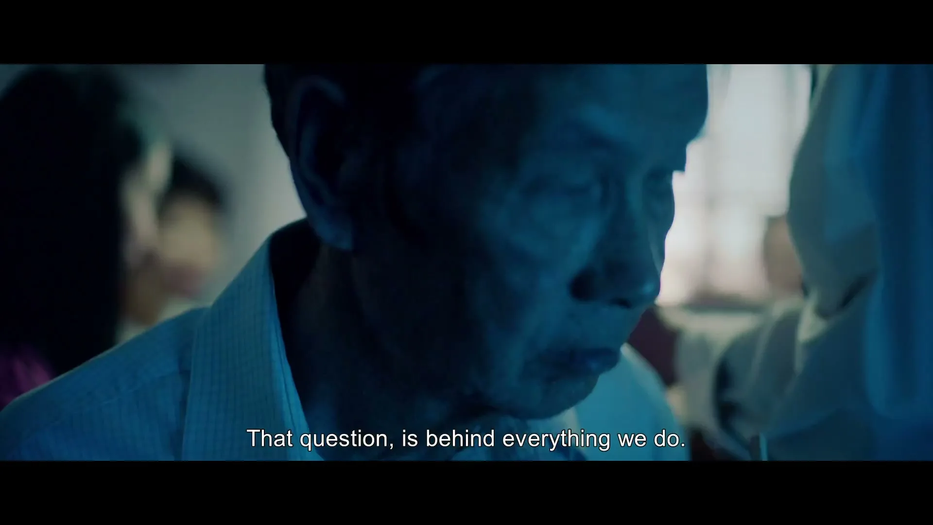 An older man with short grey hair and a white shirt looks down, appearing thoughtful, in a dimly lit room with other people in the background.