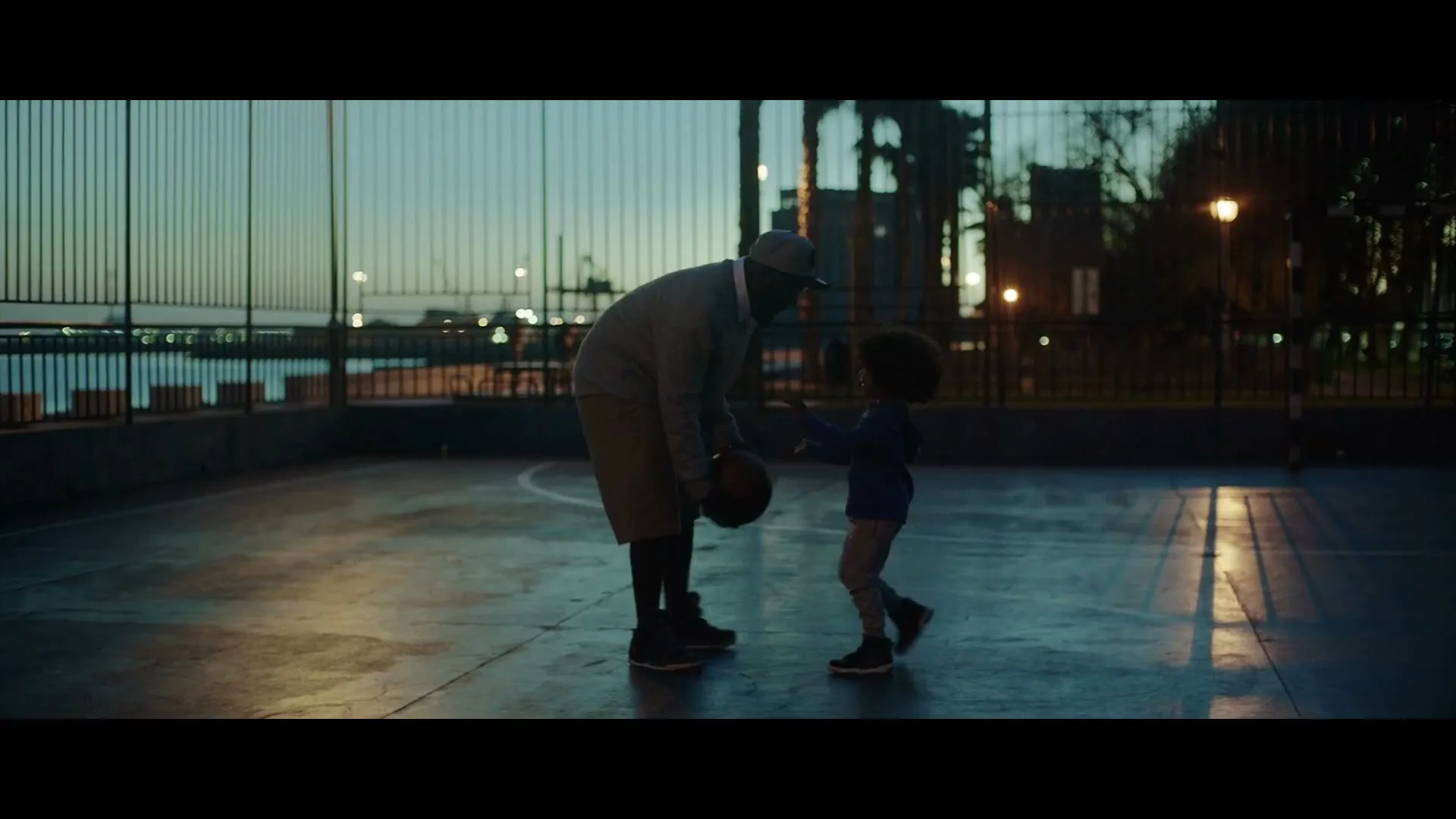 A father and his young son play basketball on an outdoor court at dusk, with a fence and water in the background.