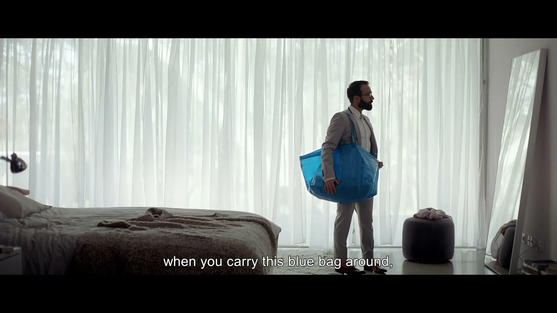 A man in a suit and red high heels carries a large blue bag around his waist, looking at himself in a mirror in a bright bedroom.