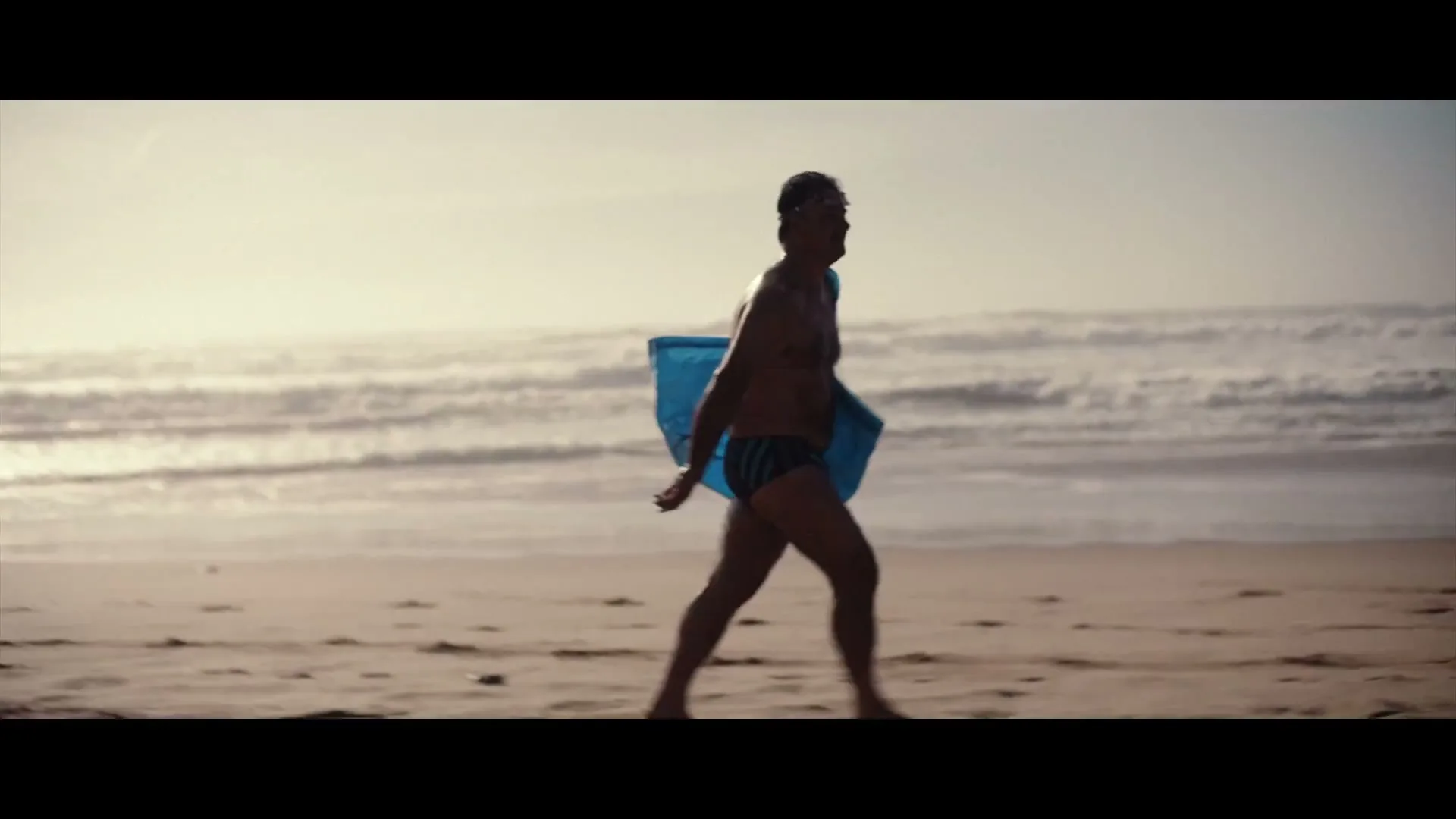 A man in his 50s, wearing a speedo and carrying a blue bag, walks along a sandy beach with waves in the background.
