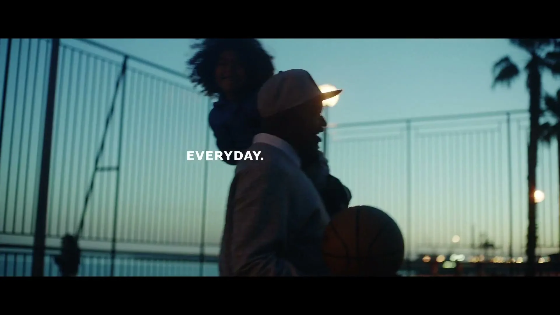 A man carries a child on his shoulders, holding a basketball, walking past a fence at dusk with palm trees in the background.