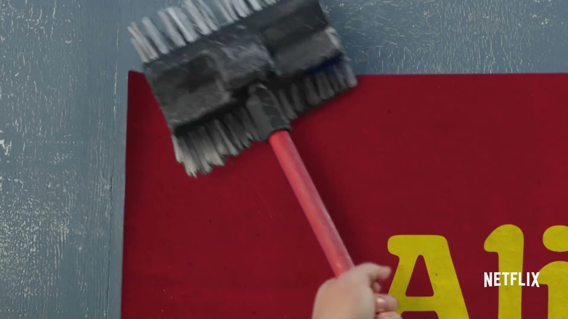A person's hand uses a broom to smooth out a large red poster with yellow text on a textured blue wall.