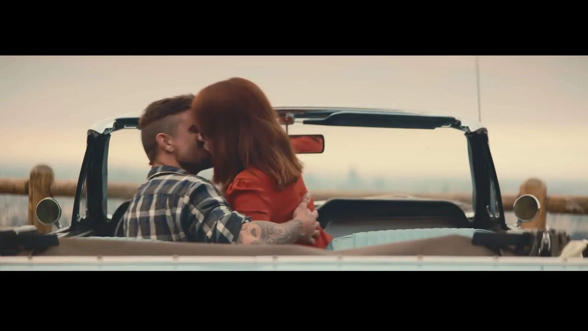A young couple, a man in a plaid shirt and a woman with red hair in a red dress, passionately kiss in a vintage convertible car, overlooking a scenic vista.