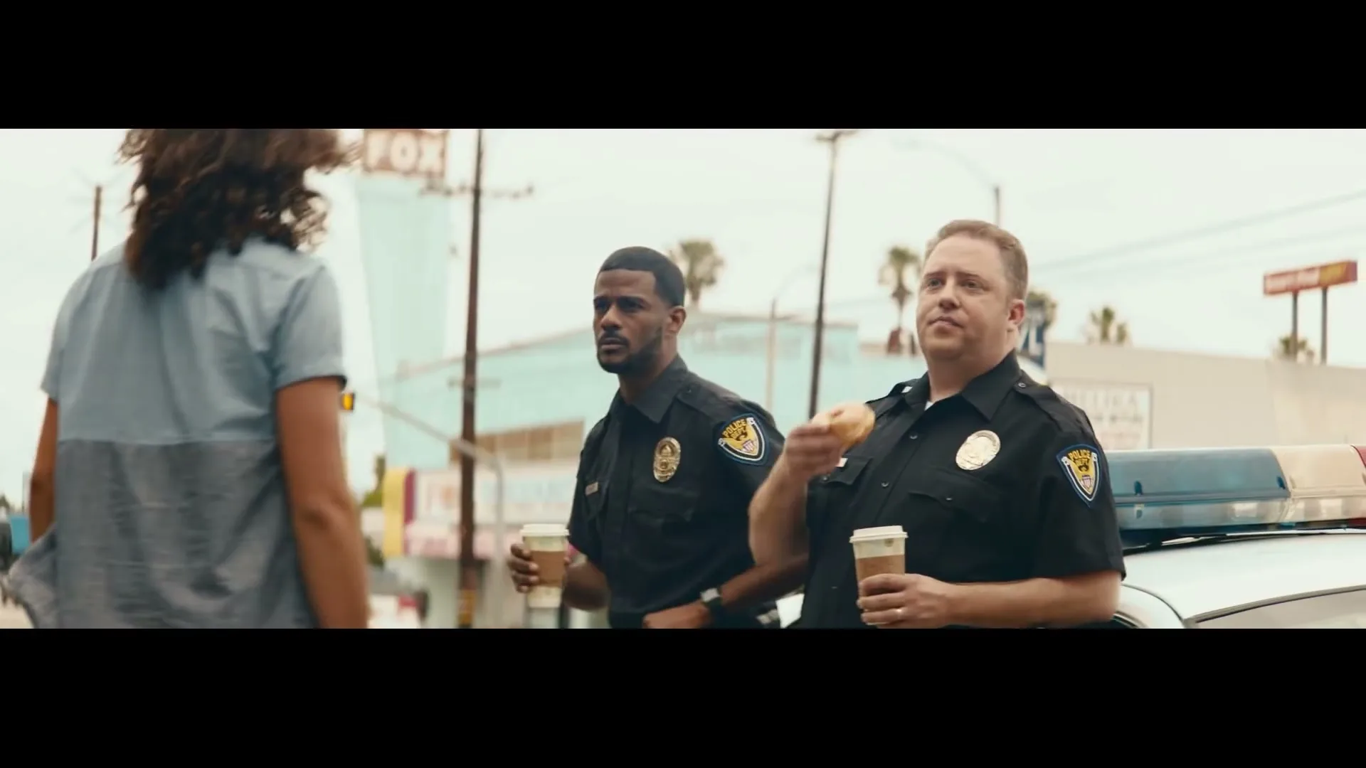 Two male police officers in uniform stand next to a patrol car, one eating a donut and drinking coffee, on a sunny street in a city.