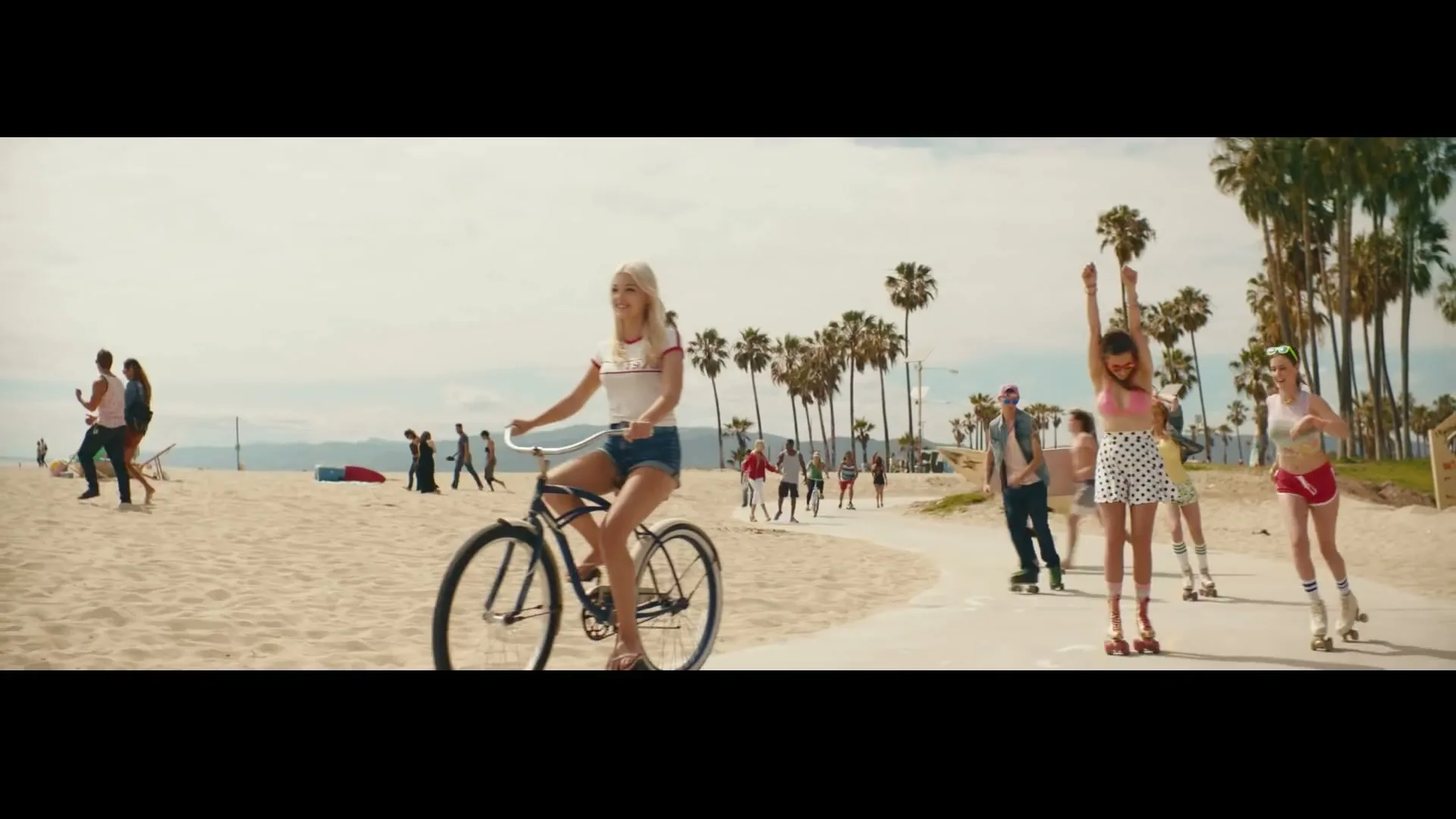 A young blonde woman rides a bicycle on a paved path next to a sandy beach, with palm trees and other people in the background.