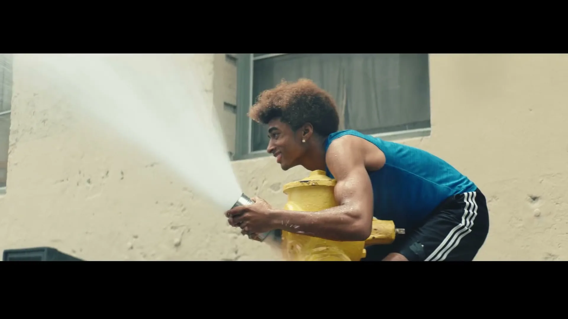 A young man with an afro, wearing a blue tank top and black shorts, smiles while spraying water from a fire hydrant on a city street.
