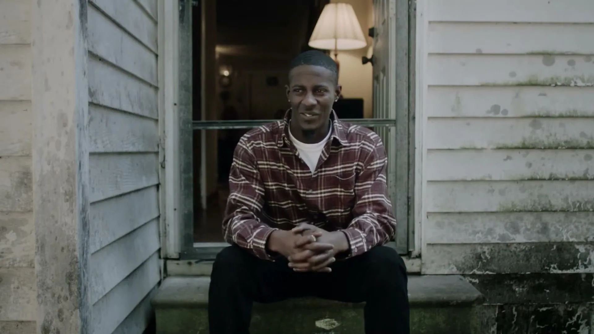 A young Black man in a plaid shirt sits on the porch of a weathered house, looking directly at the camera with a slight smile.