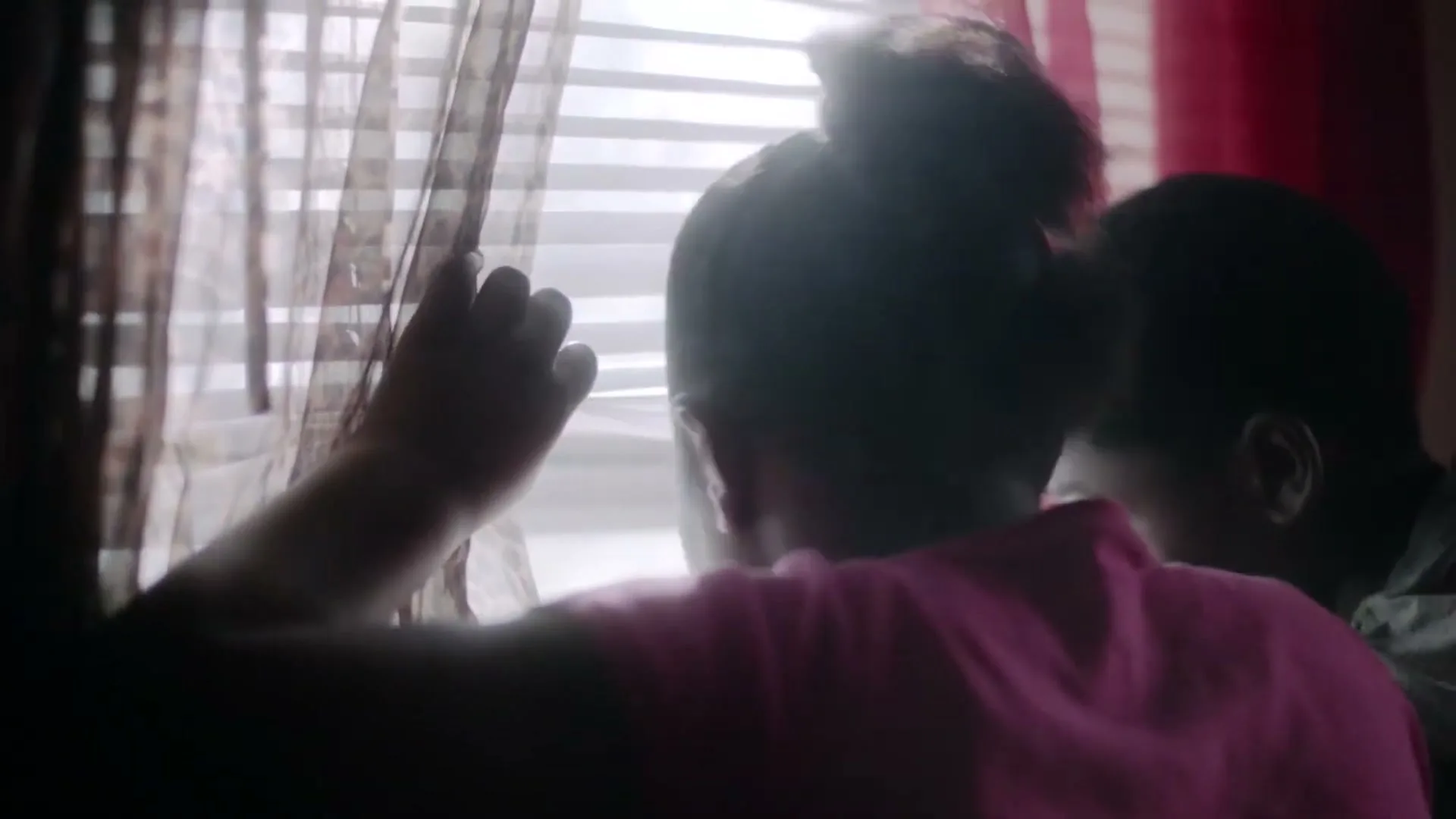 Two young children, a girl in a pink shirt and a boy, look out a window with blinds and sheer curtains, their backs to the camera.