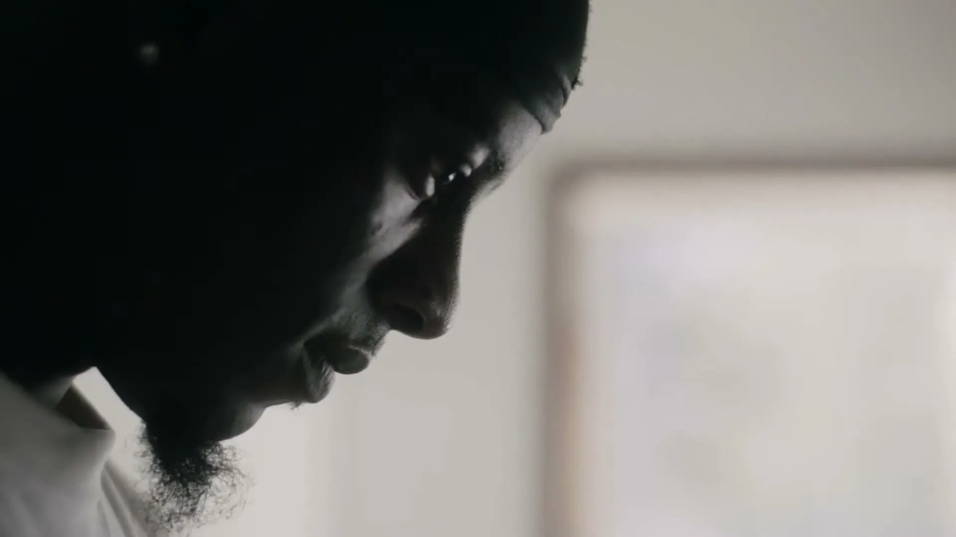 A young Black man in a durag and white shirt is seen in profile, looking down with a serious expression in a dimly lit room.