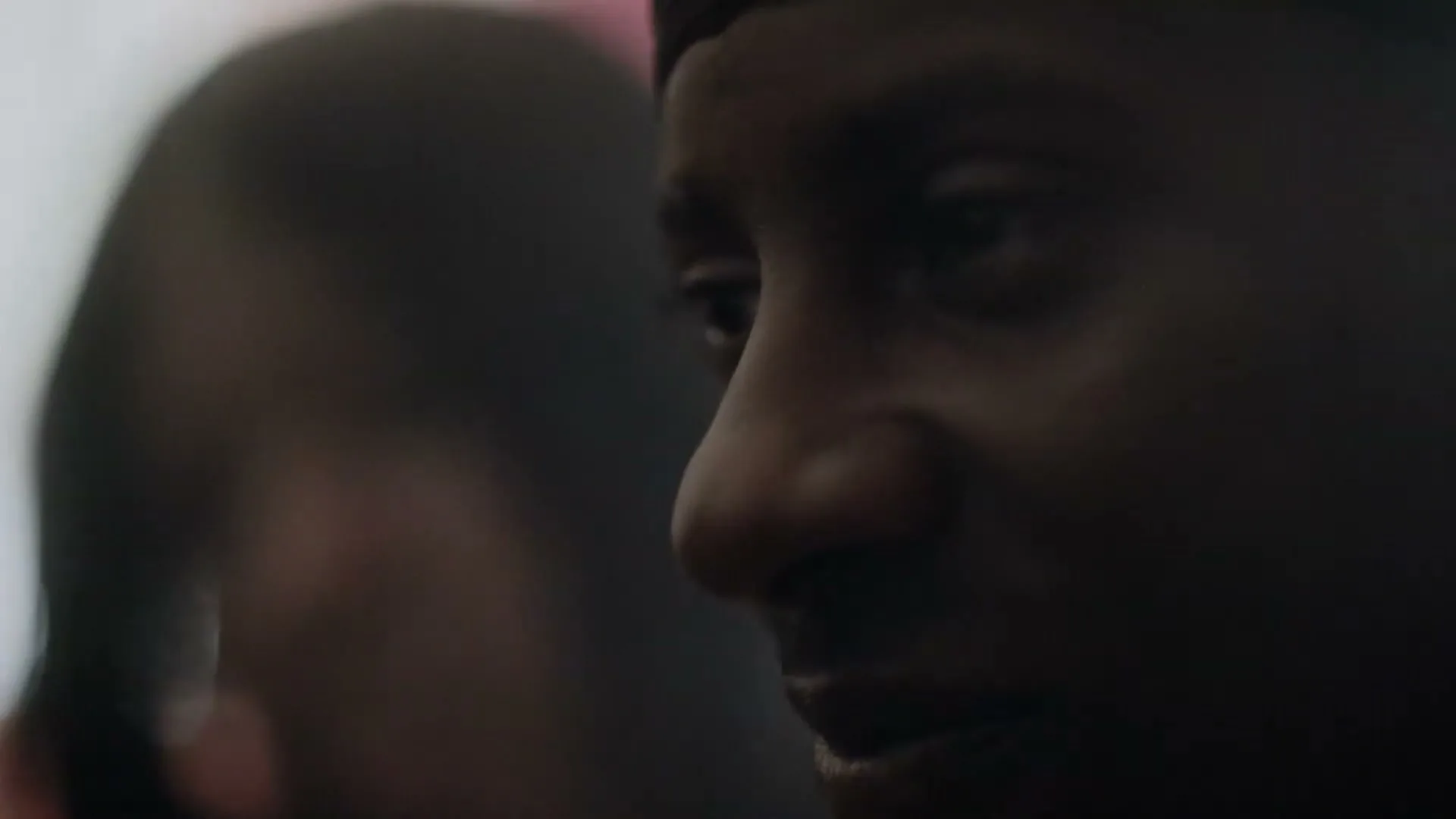 A man with a dark durag looks off-camera, his face partially obscured by shadow, as another person is out of focus in the background.