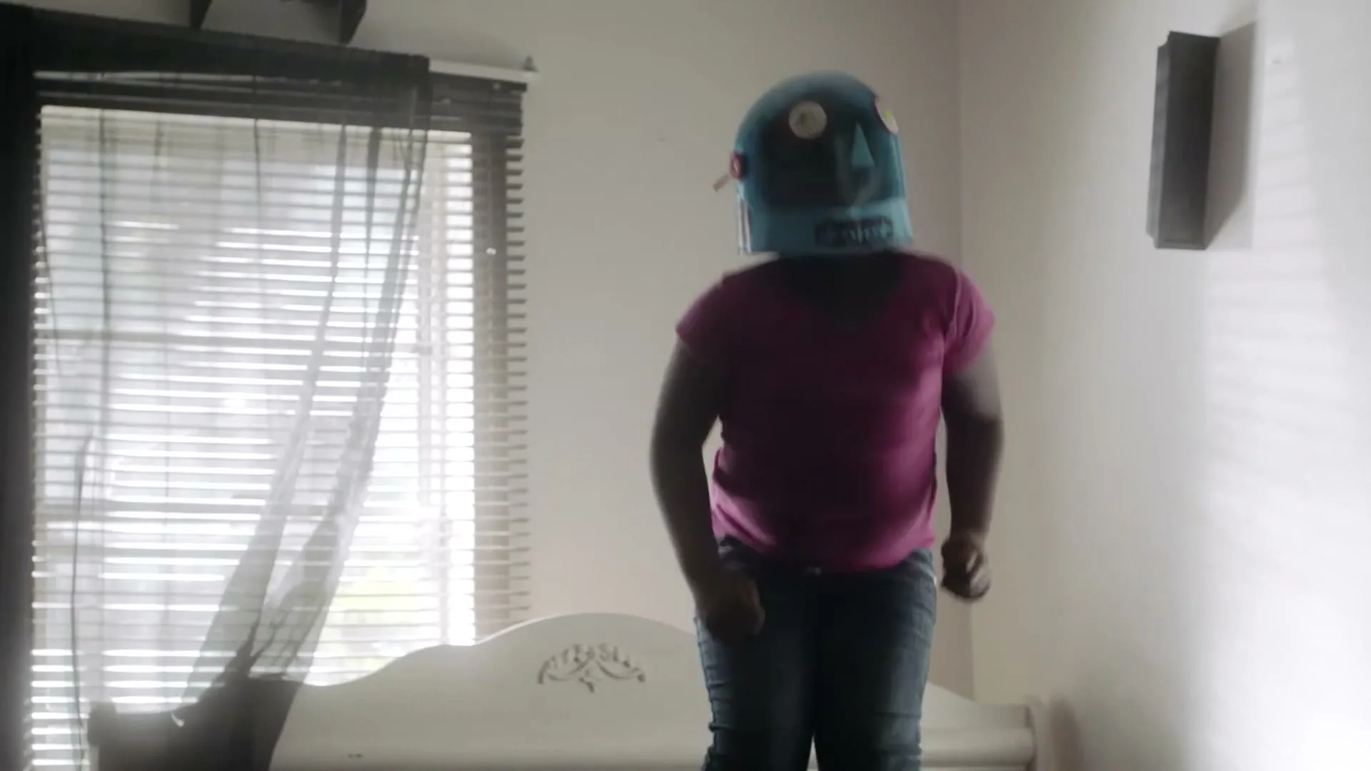 A child wearing a blue robot helmet and pink shirt jumps on a bed in a brightly lit bedroom with blinds on the window.