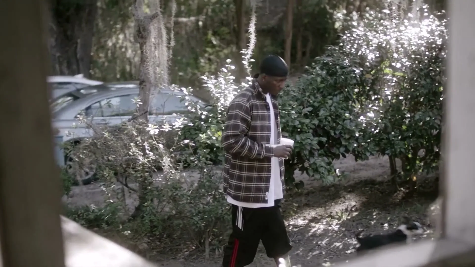 A man in a plaid shirt and black shorts walks with a cup in his hand, with cars and trees in the background.