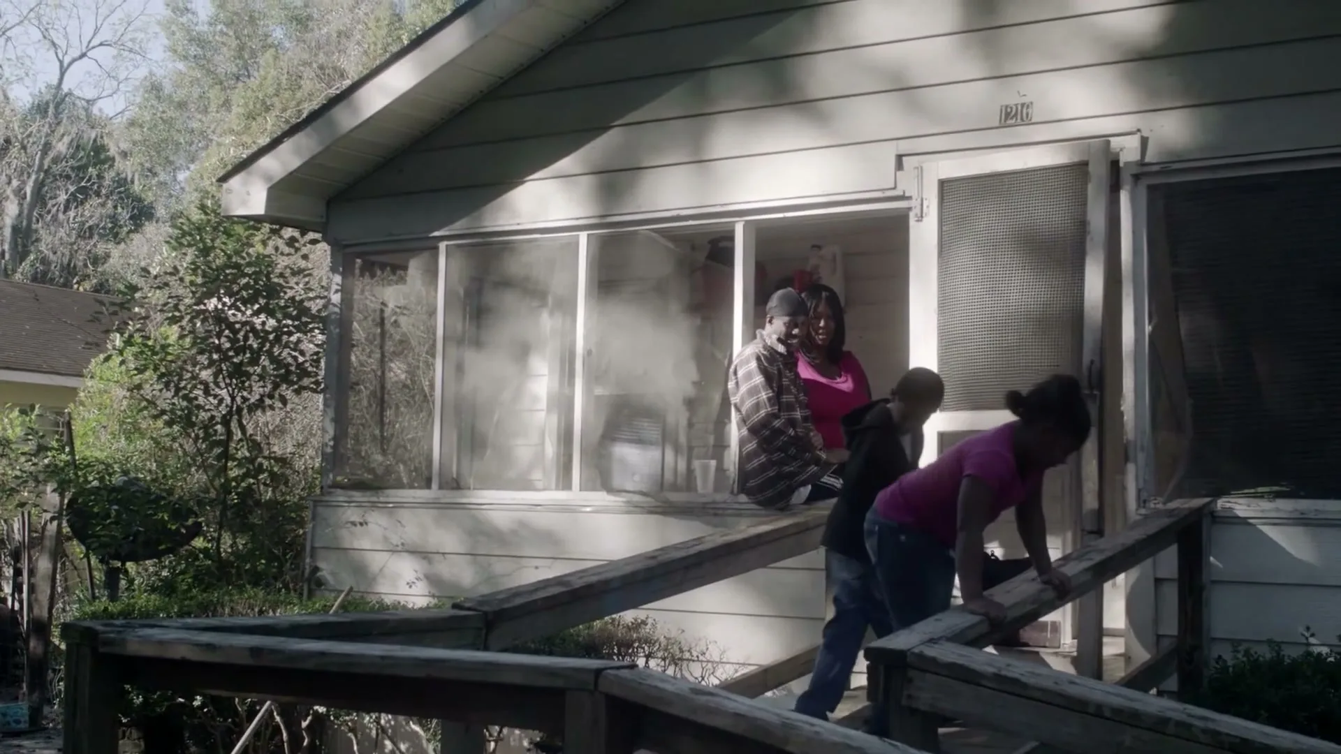 A family of four, including two adults and two children, are seen on the porch of a house, with smoke emanating from inside the house.
