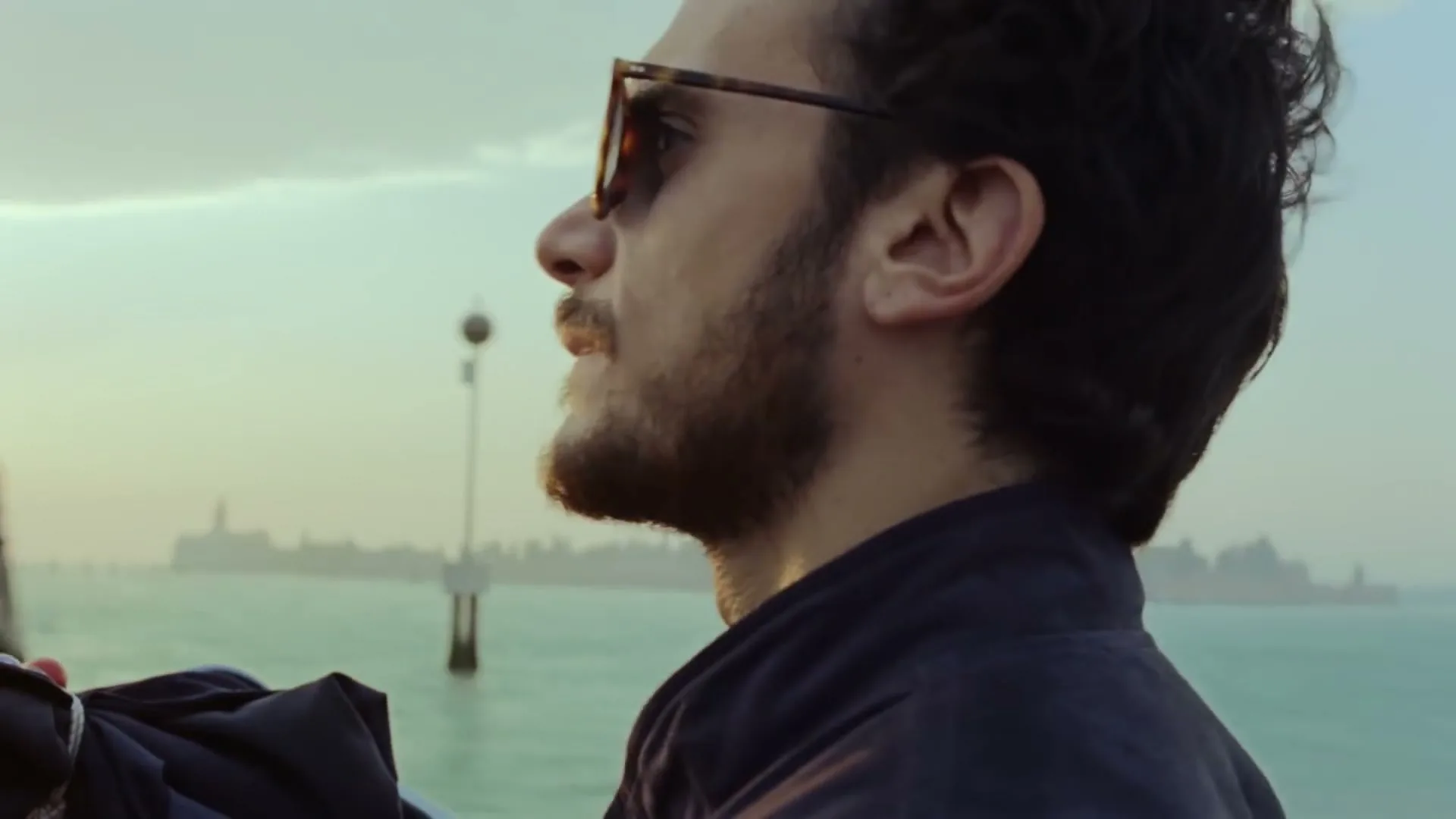 A man with sunglasses and a beard looks out over the water at a distant city skyline during golden hour.