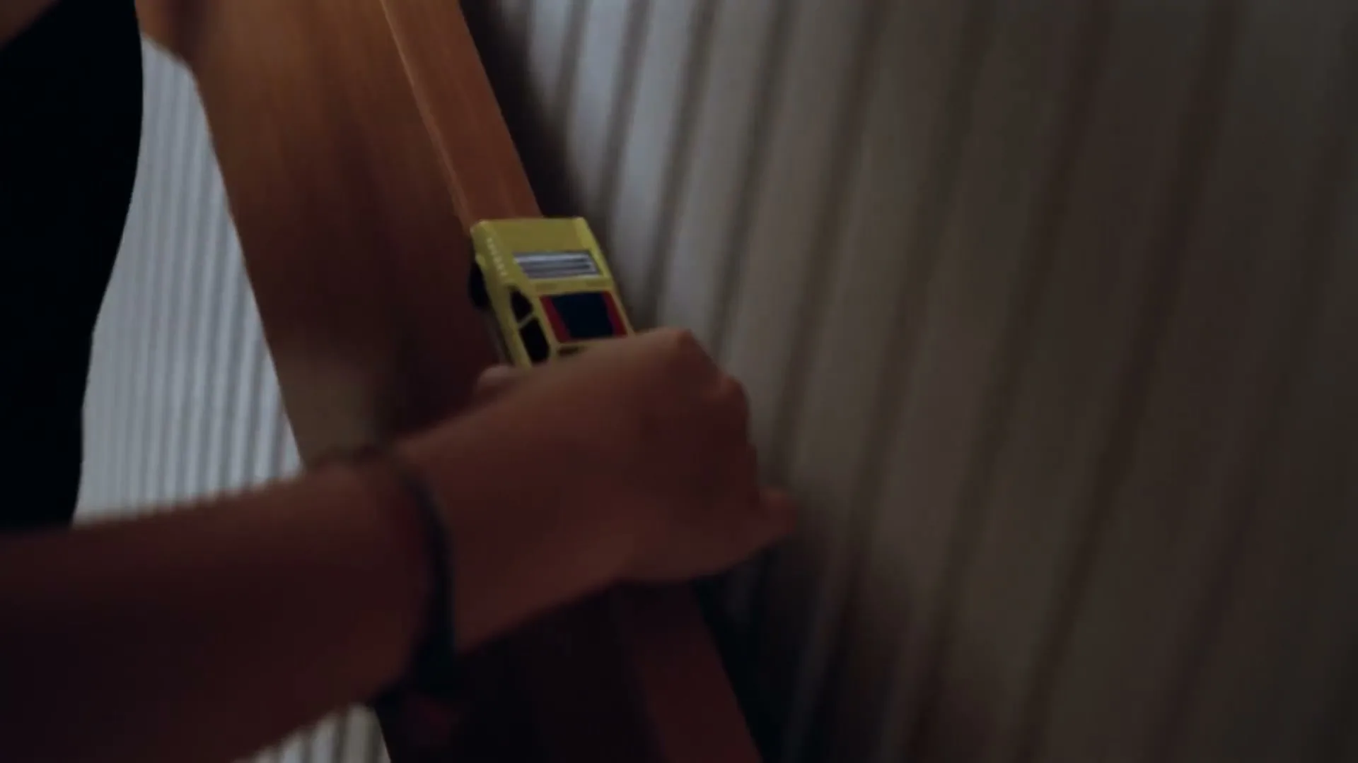 A person's hands are seen playing with a yellow toy car on a wooden surface next to a ribbed wall.