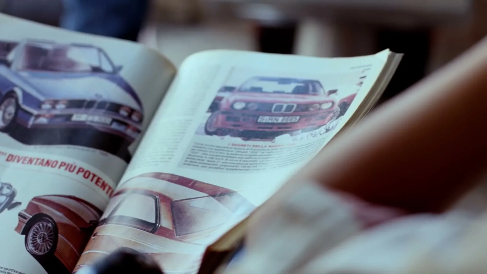 A person holds open a magazine featuring a red BMW car on the right page, with other car images on the left page, in an interior setting.