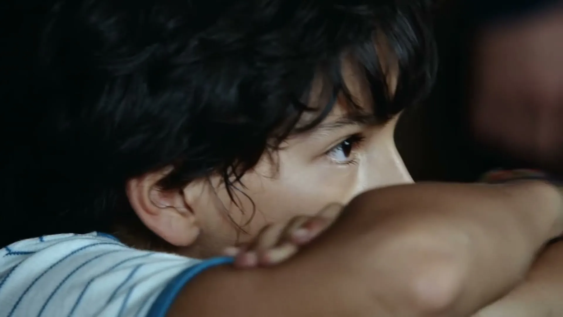 A young boy with dark, curly hair looks intently to the right, resting his chin on his crossed arms, in a medium closeup shot.