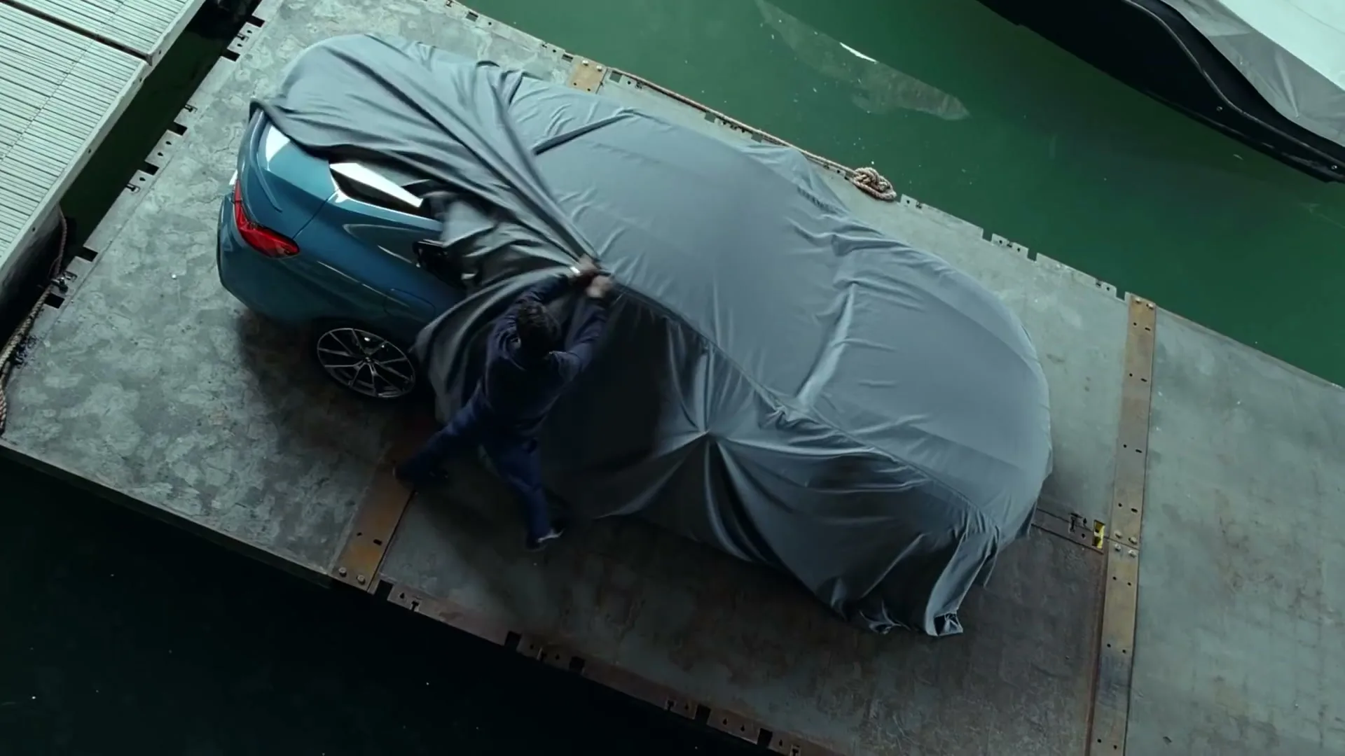 A man in a dark jacket pulls a grey car cover off a blue car on a floating dock next to green water.