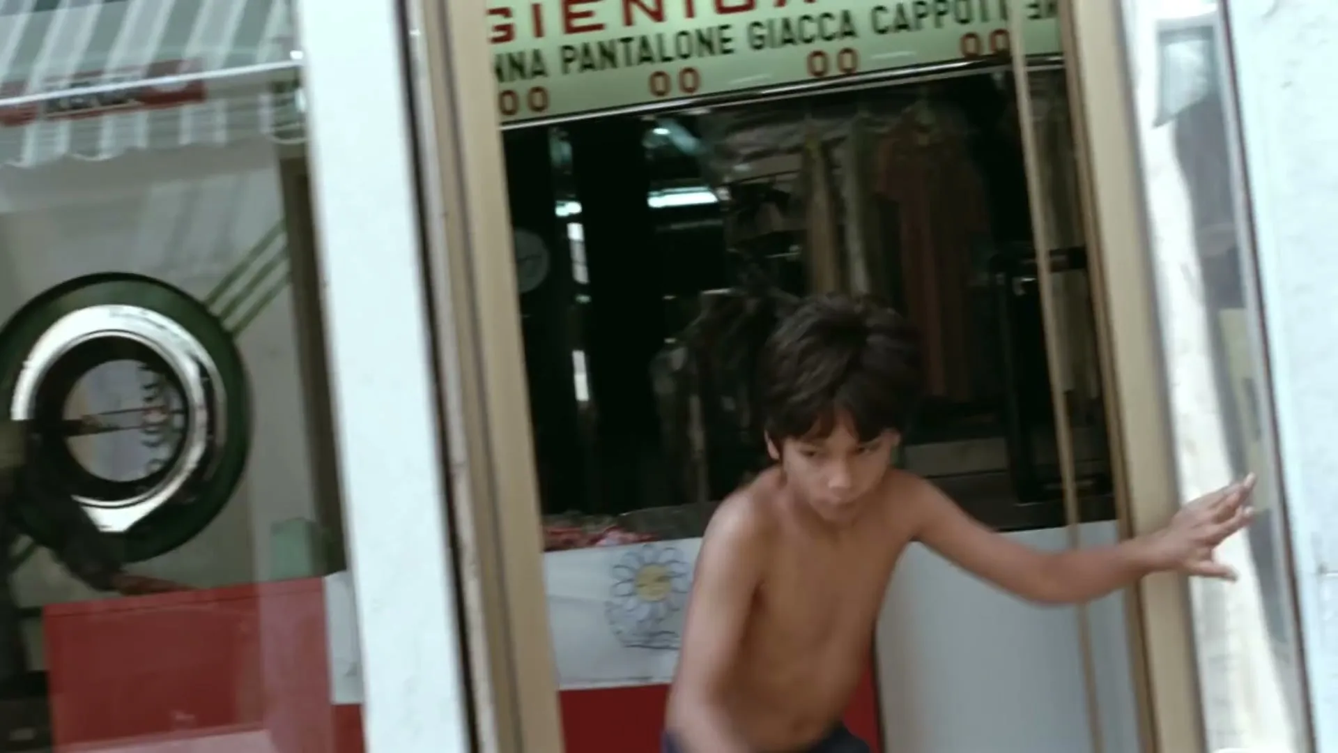 A shirtless young boy runs out of a laundromat, with a woman visible in the background through a window, in a bright, sunny setting.