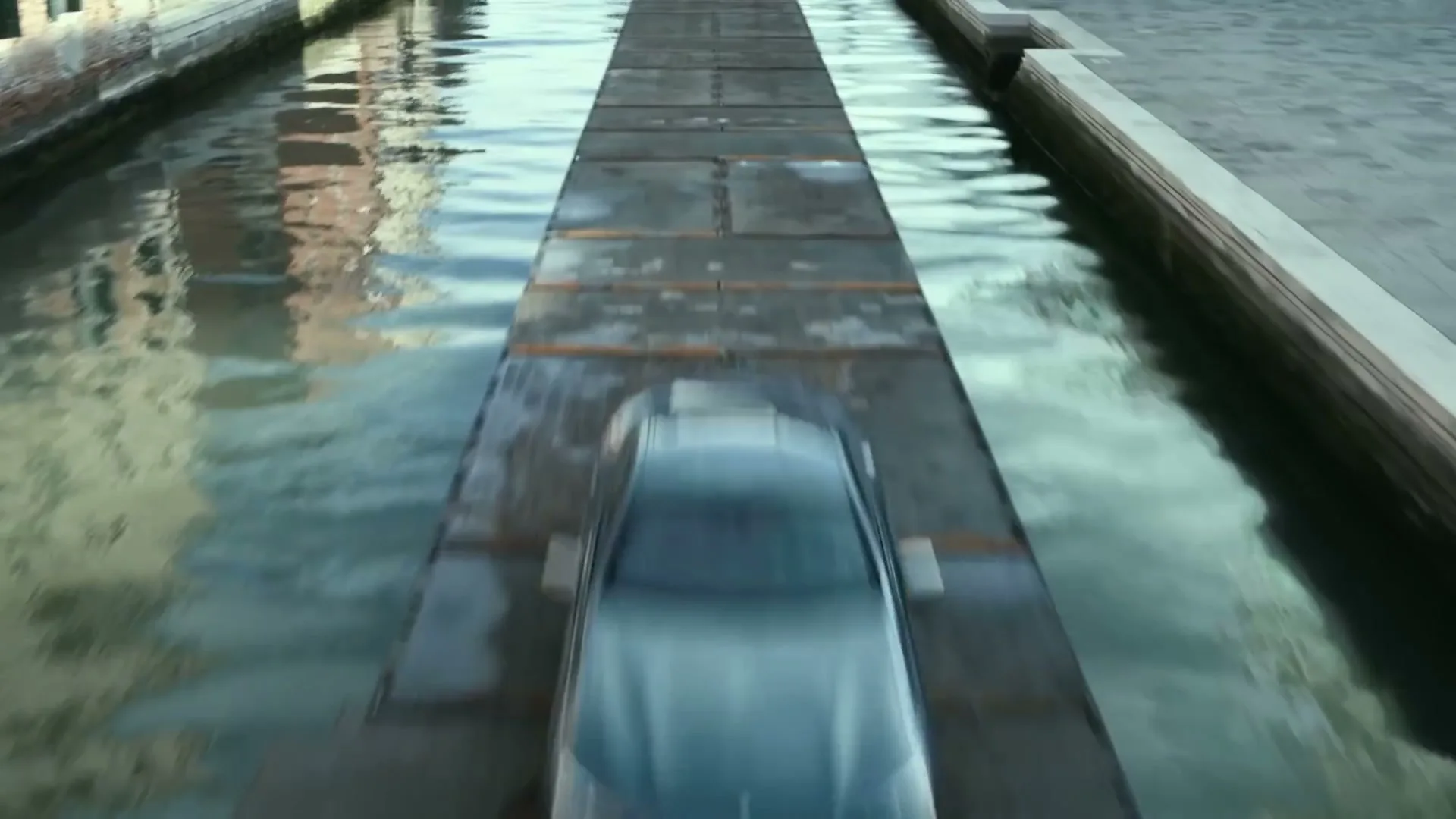 A dark grey BMW 8 Series drives away on a narrow, temporary concrete path over a canal in a city with no other cars.
