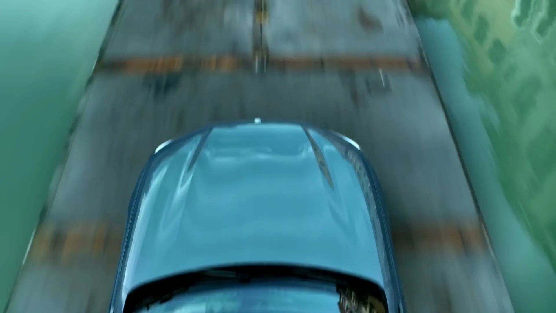 A blue BMW 8 Series drives quickly over a narrow concrete bridge, with water on both sides, in an empty city.