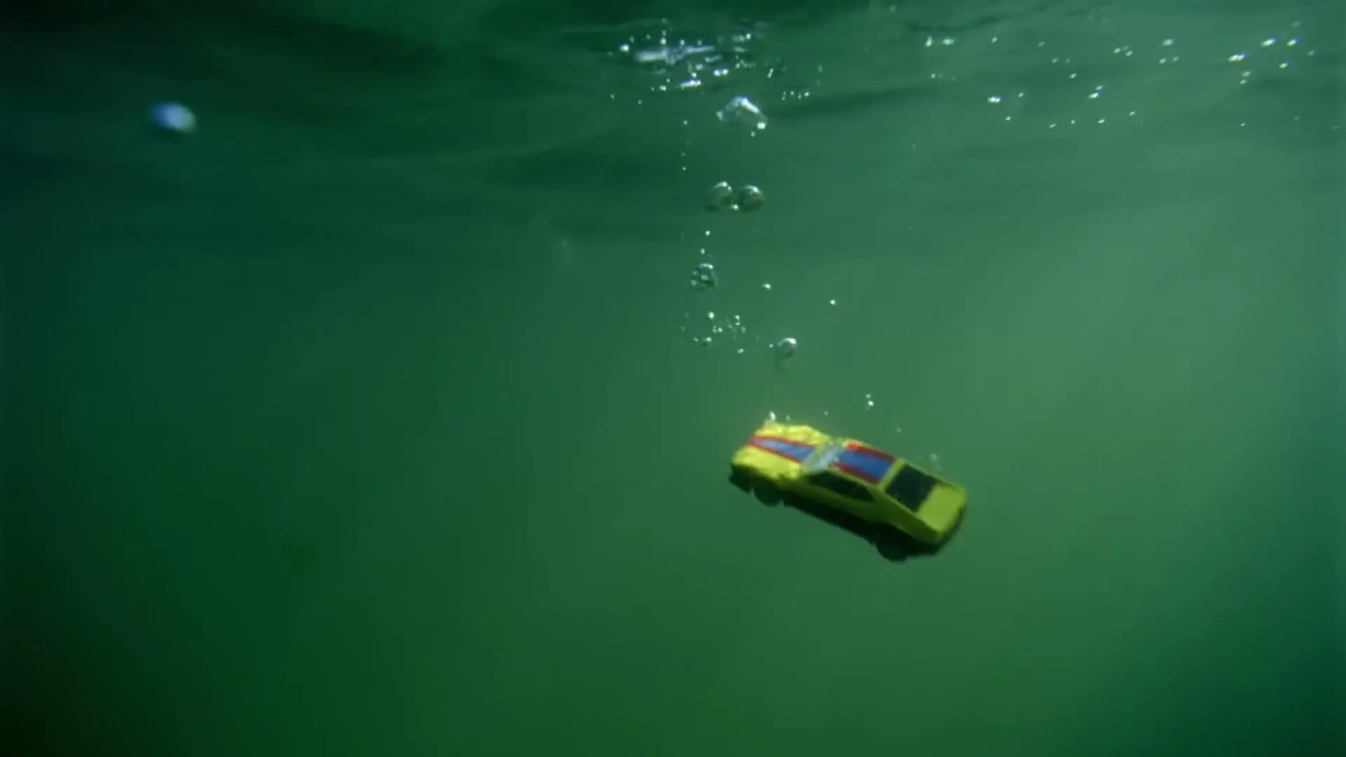 A yellow toy car with red and blue accents sinks slowly in murky green water, with bubbles rising to the surface.