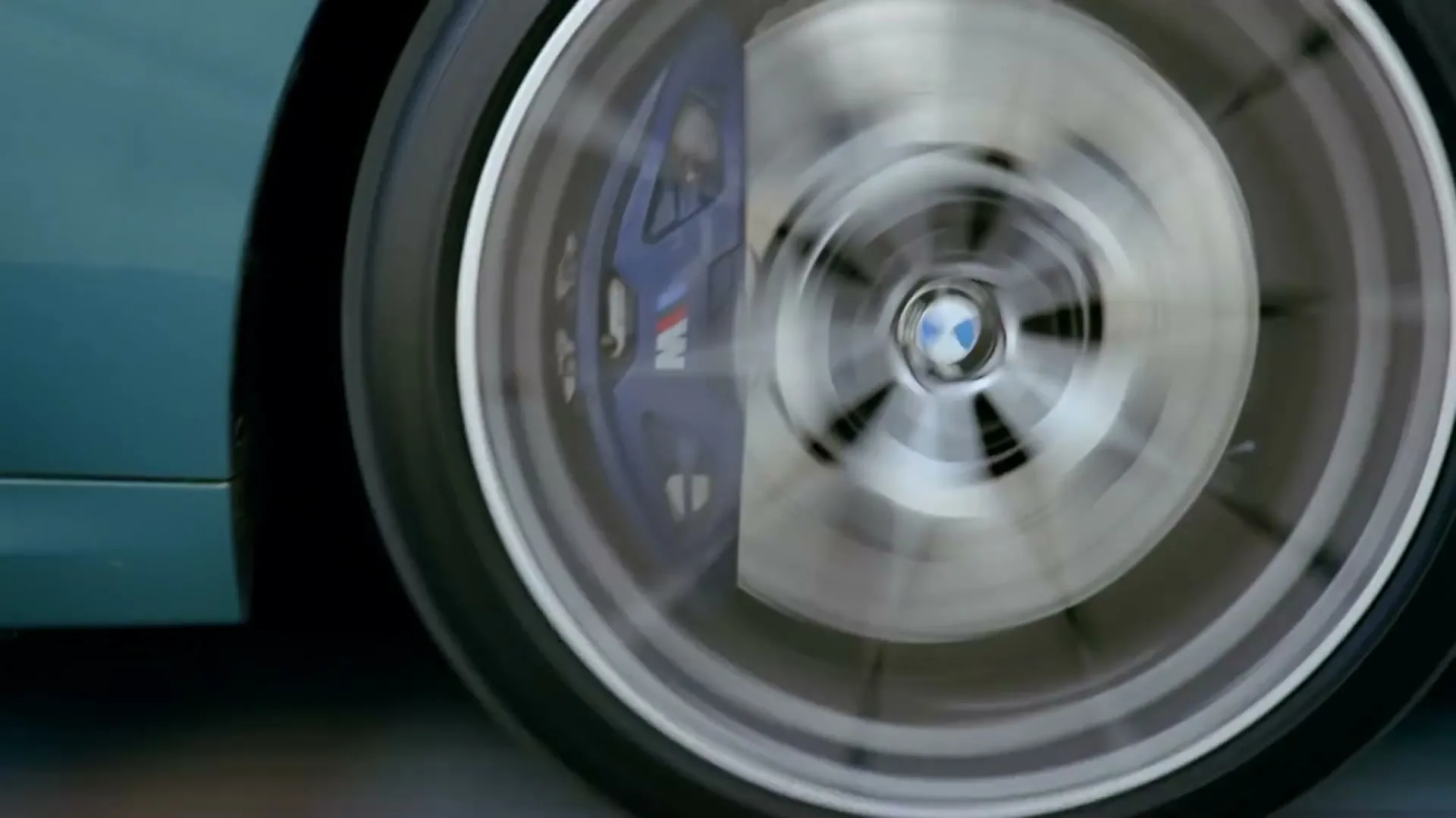 A close-up shot of a BMW car wheel with blue brake calipers, spinning rapidly, as the car drives on a road.