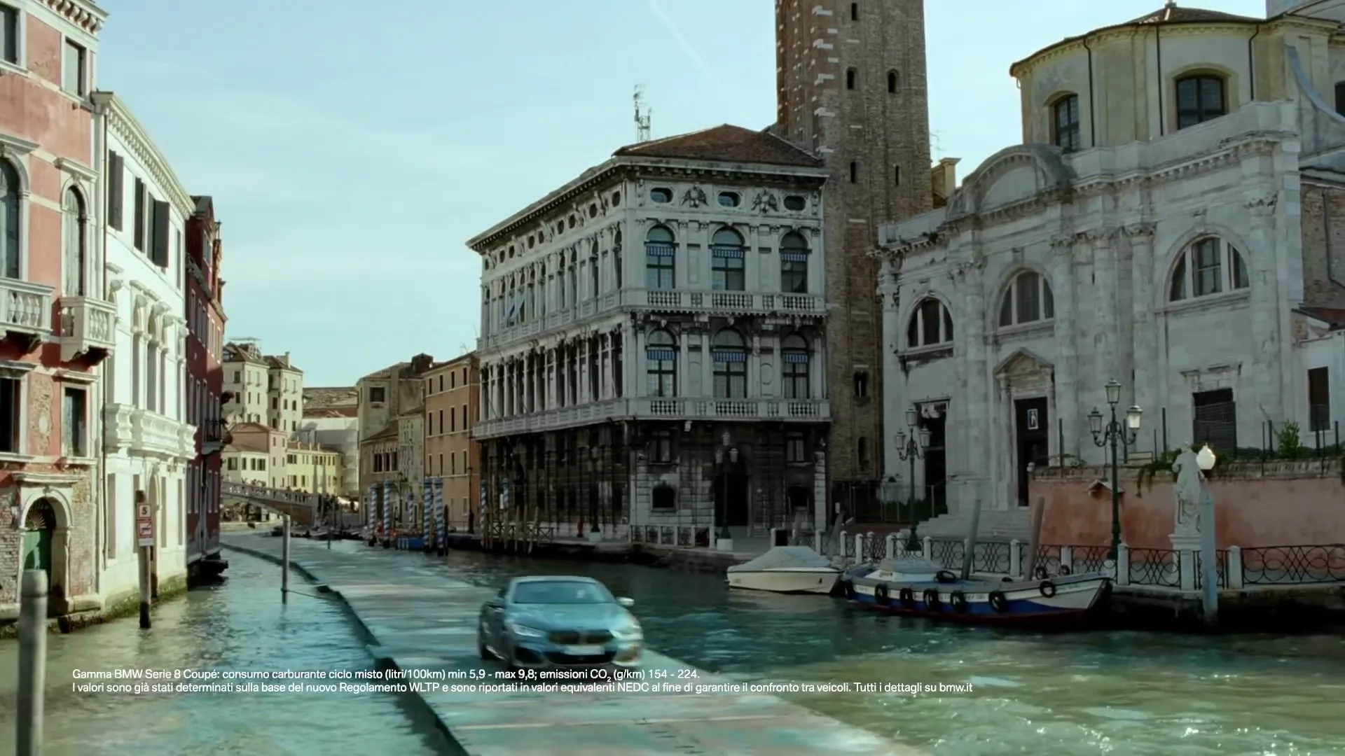 A blue BMW 8 Series Coupe drives on a narrow, wet stone path next to a canal in Venice, Italy, surrounded by historic buildings under a bright sky.