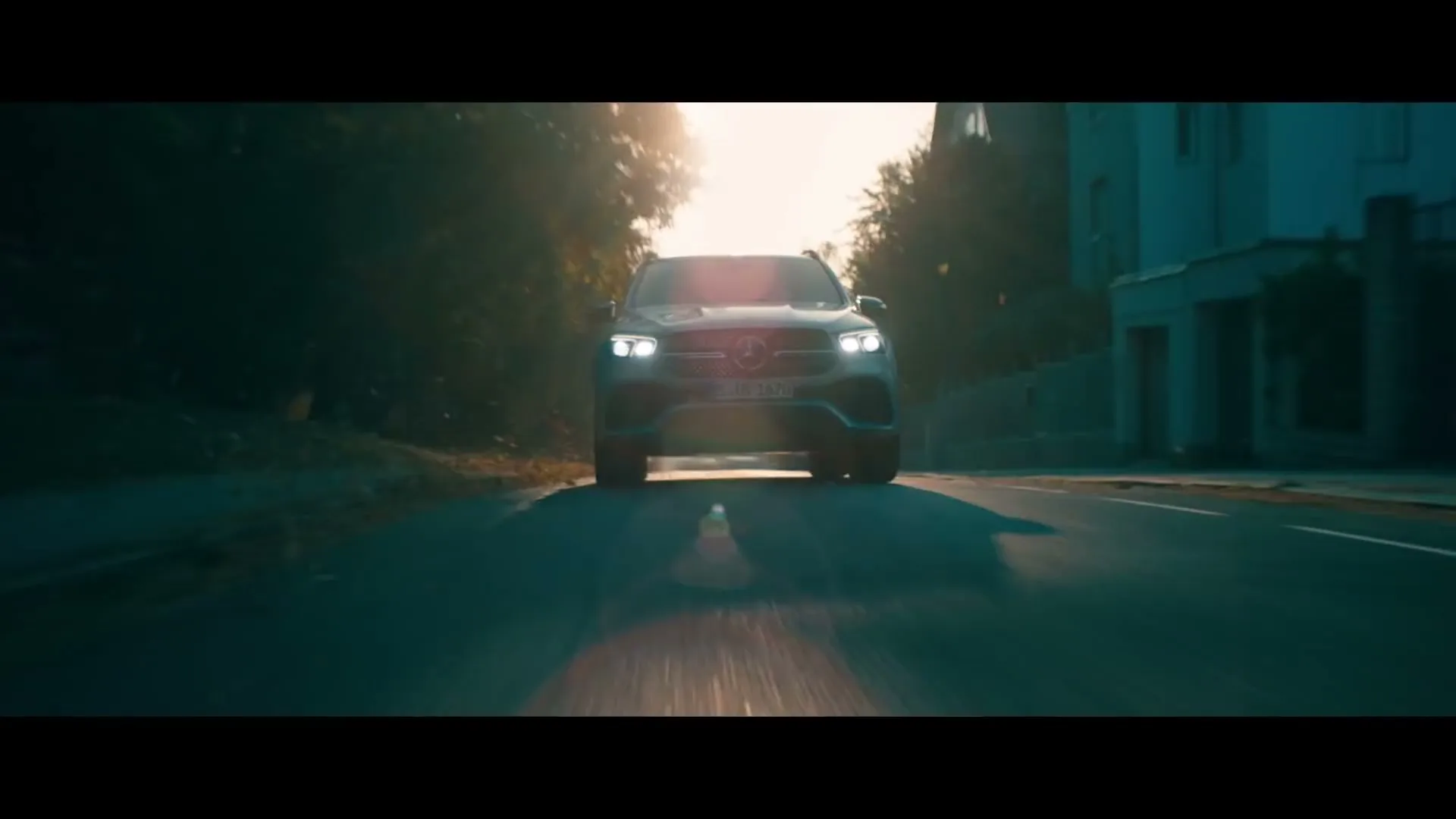 A dark Mercedes-Benz SUV drives towards the camera on a tree-lined street during golden hour, with bright headlights on.