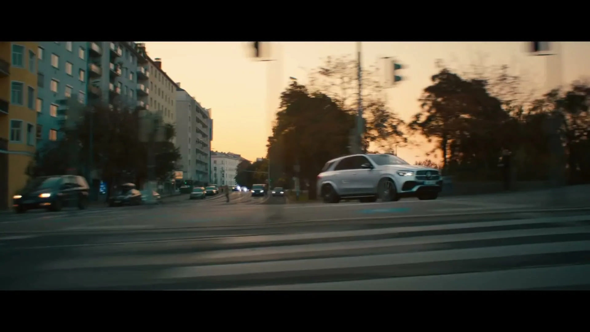 A silver Mercedes-Benz SUV drives quickly across a city intersection at sunset, with buildings and trees in the background.