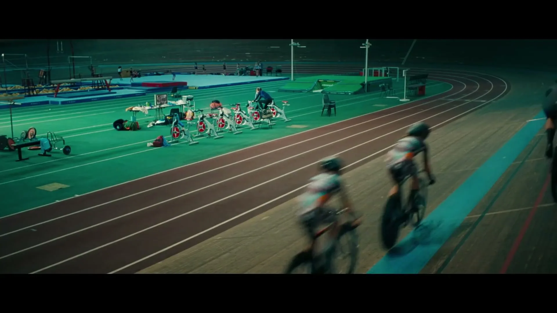 Cyclists race on a track in a velodrome, while other athletes train in the background, in a wide shot.