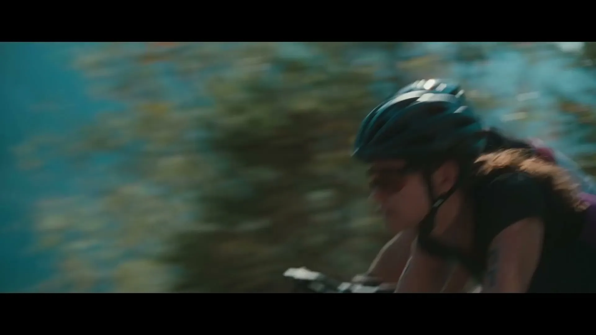 A female cyclist in a black and purple cycling suit and helmet rides a bicycle on a sunny day, with another cyclist behind her.