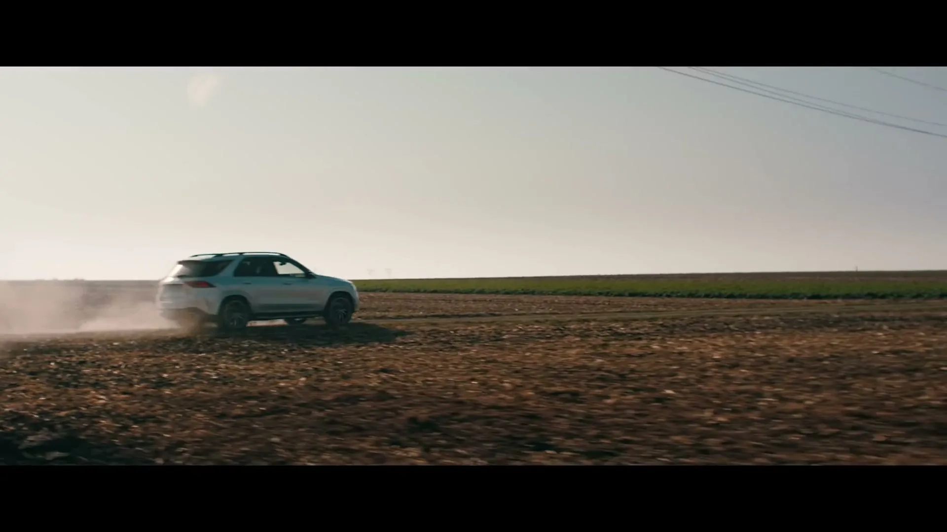 A white Mercedes-Benz SUV drives through a dusty, dry field, kicking up a cloud of dust behind it under a clear sky.