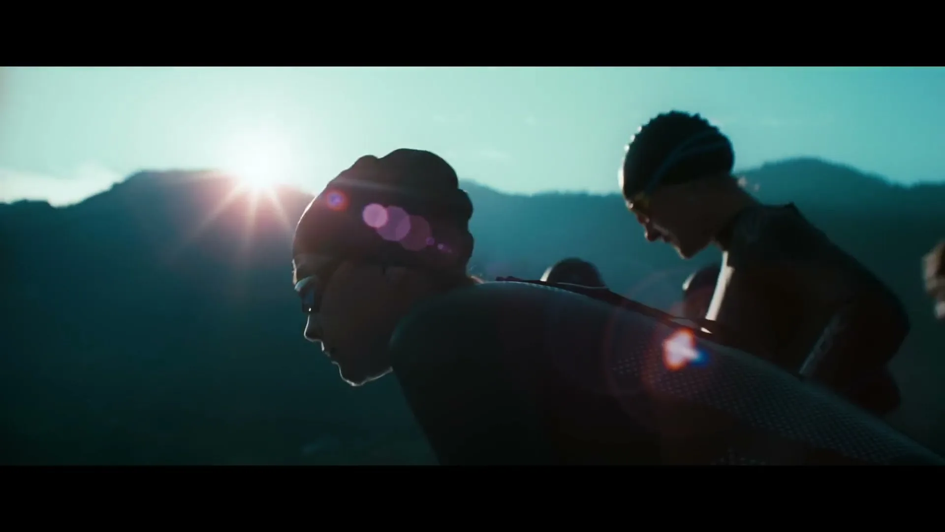 Two female triathletes in wetsuits and swim caps prepare to dive into water at dawn, silhouetted against a mountain range.