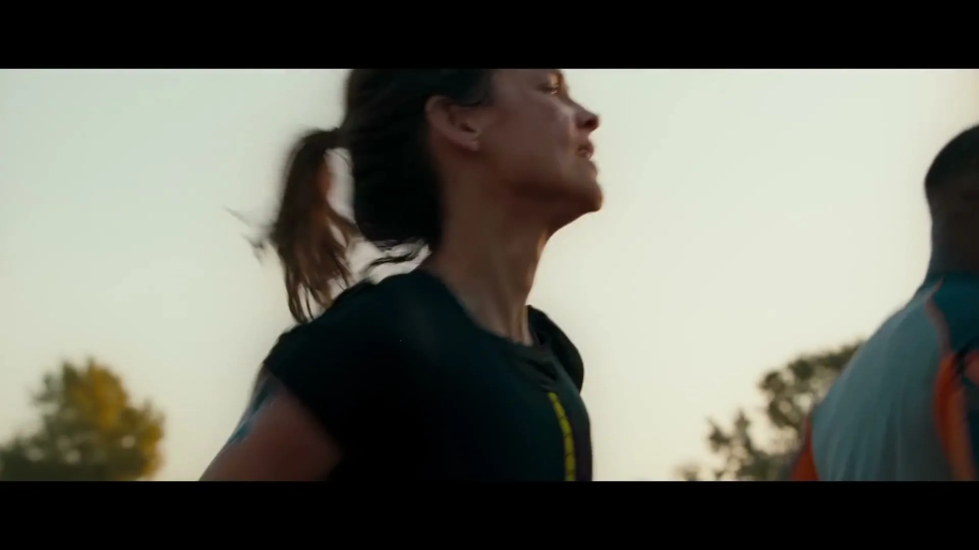 A female runner with a ponytail and black top runs on a track, with a male runner partially visible, against a bright sky.