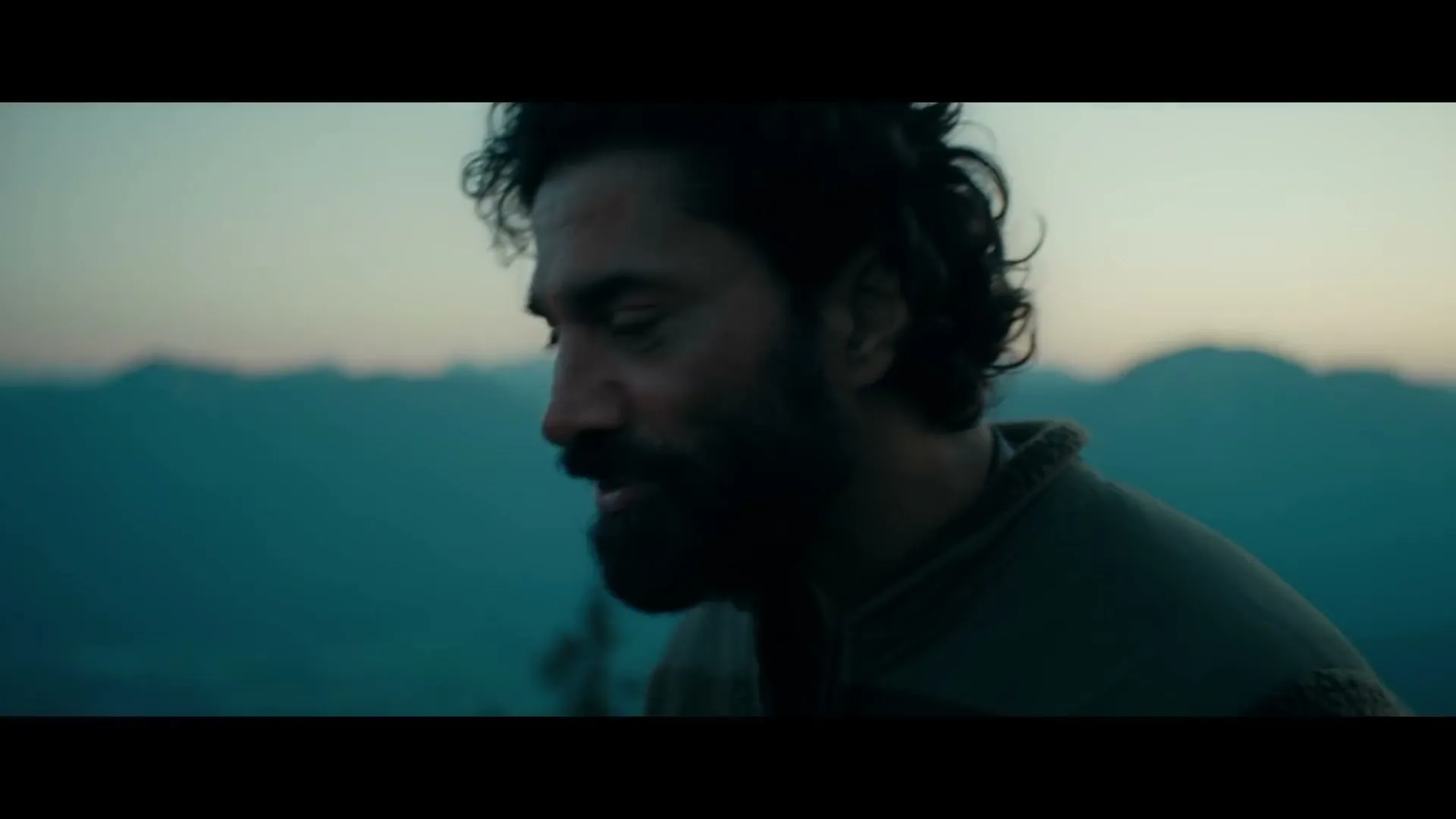 A man with a beard and dark, curly hair looks down, with mountains in the background at dusk.