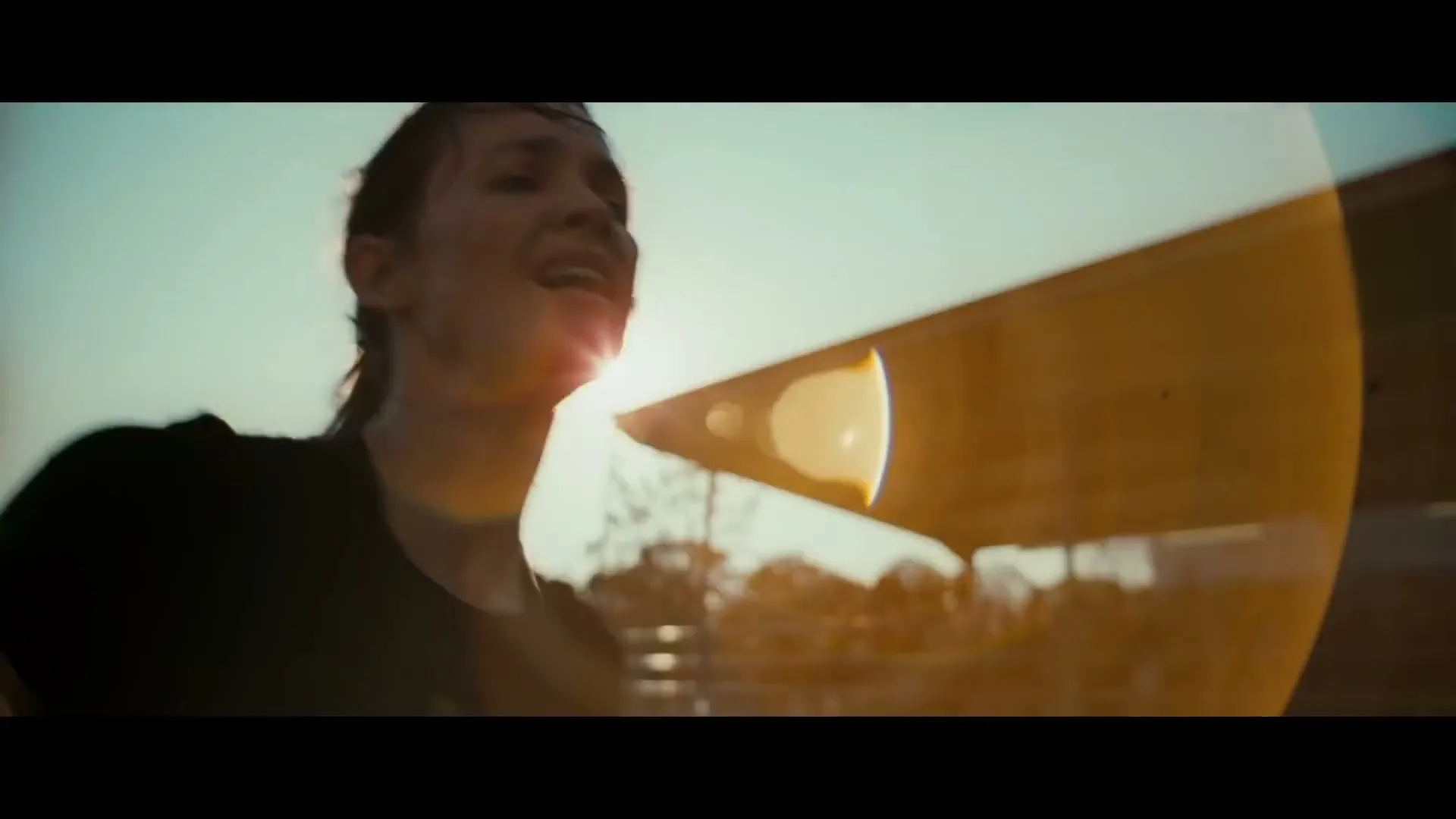 A female athlete, exhausted and sweating, runs on a track with a stadium stand in the background, bathed in golden hour sunlight.