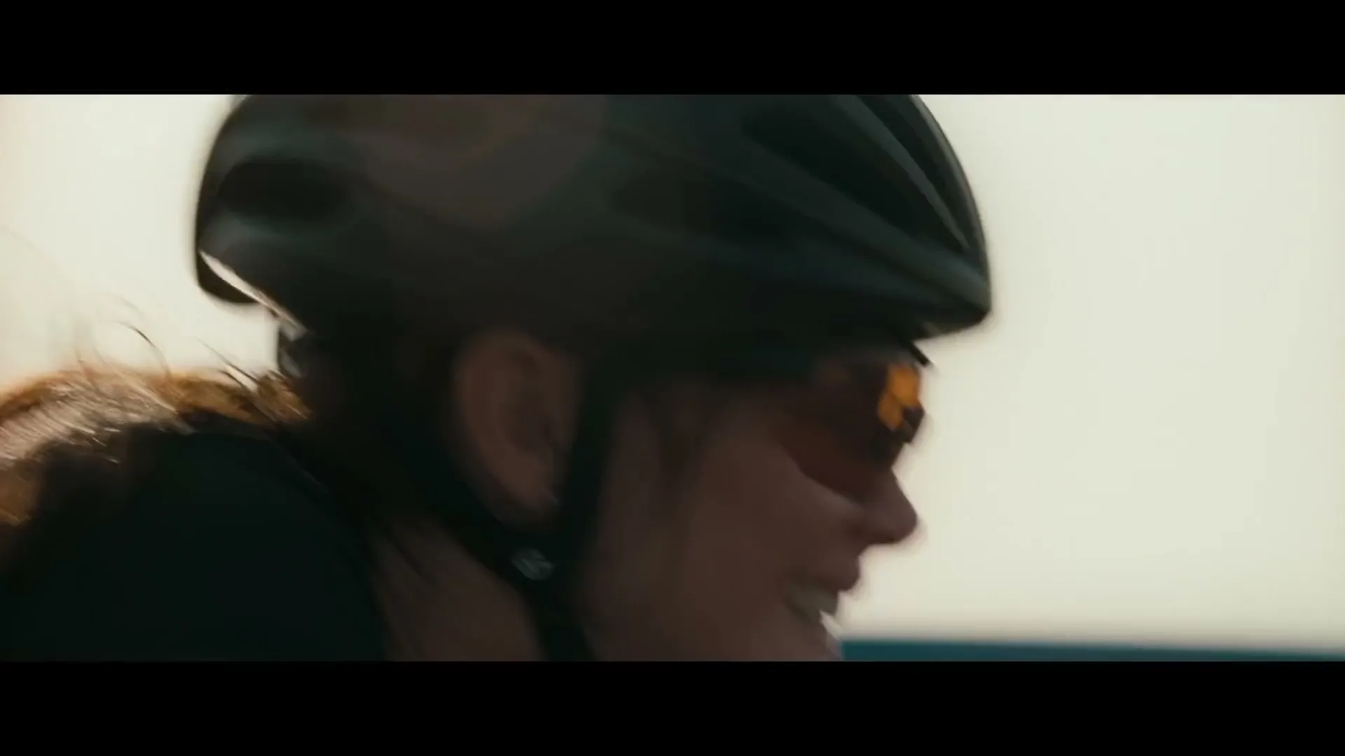 A female cyclist in a helmet and sunglasses smiles while riding a bike outdoors on a sunny day.