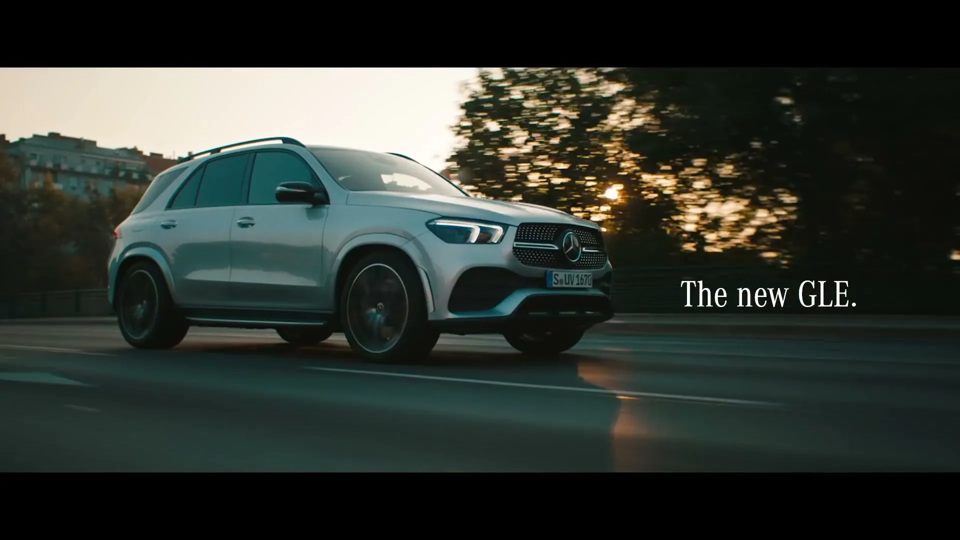 A silver Mercedes-Benz GLE SUV drives on a road at golden hour, with the sun setting behind it.