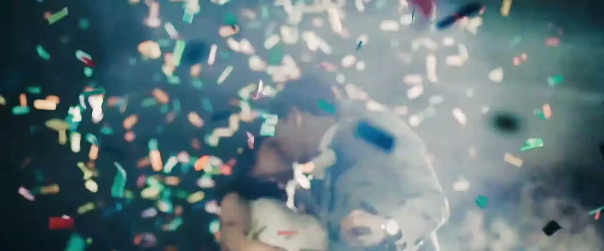 A newlywed couple, a man in a suit and a woman in a white dress, kiss as colorful confetti falls around them in a celebratory moment.